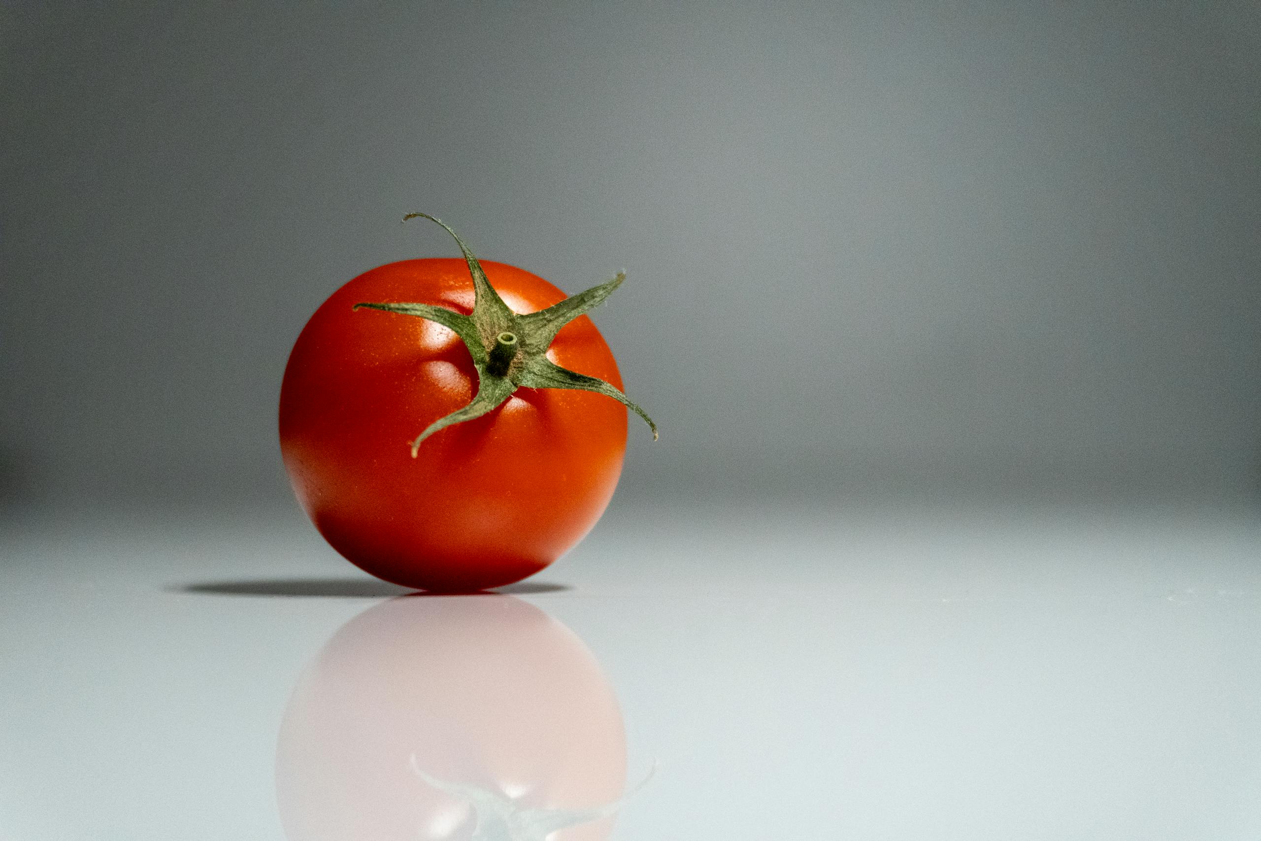 A solitary fresh tomato on a reflective surface, showcasing vibrant red color and traditional green stem.