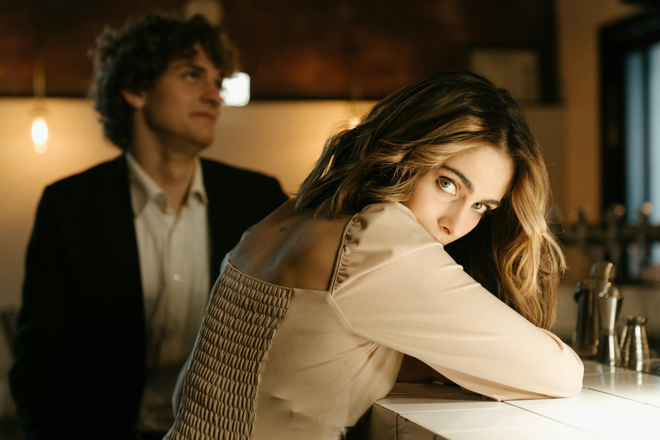 A stylish man and woman share a moment at a bar, with warm ambient lighting.