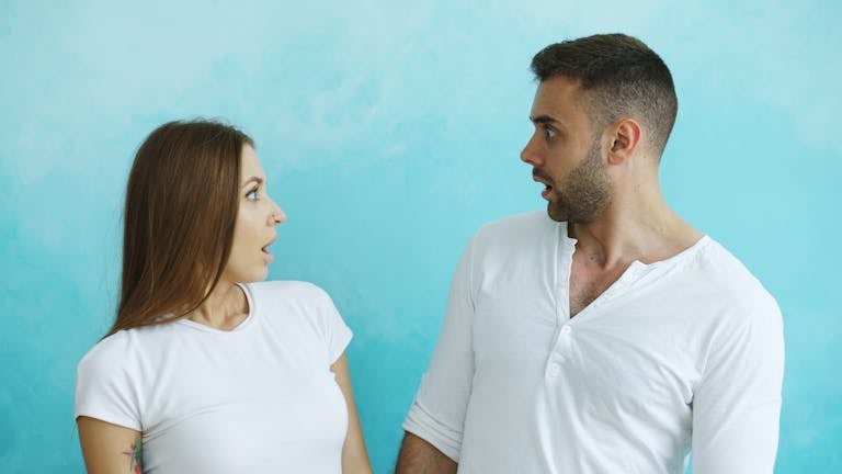 A surprised man and woman facing each other with expressions of shock against a blue background.