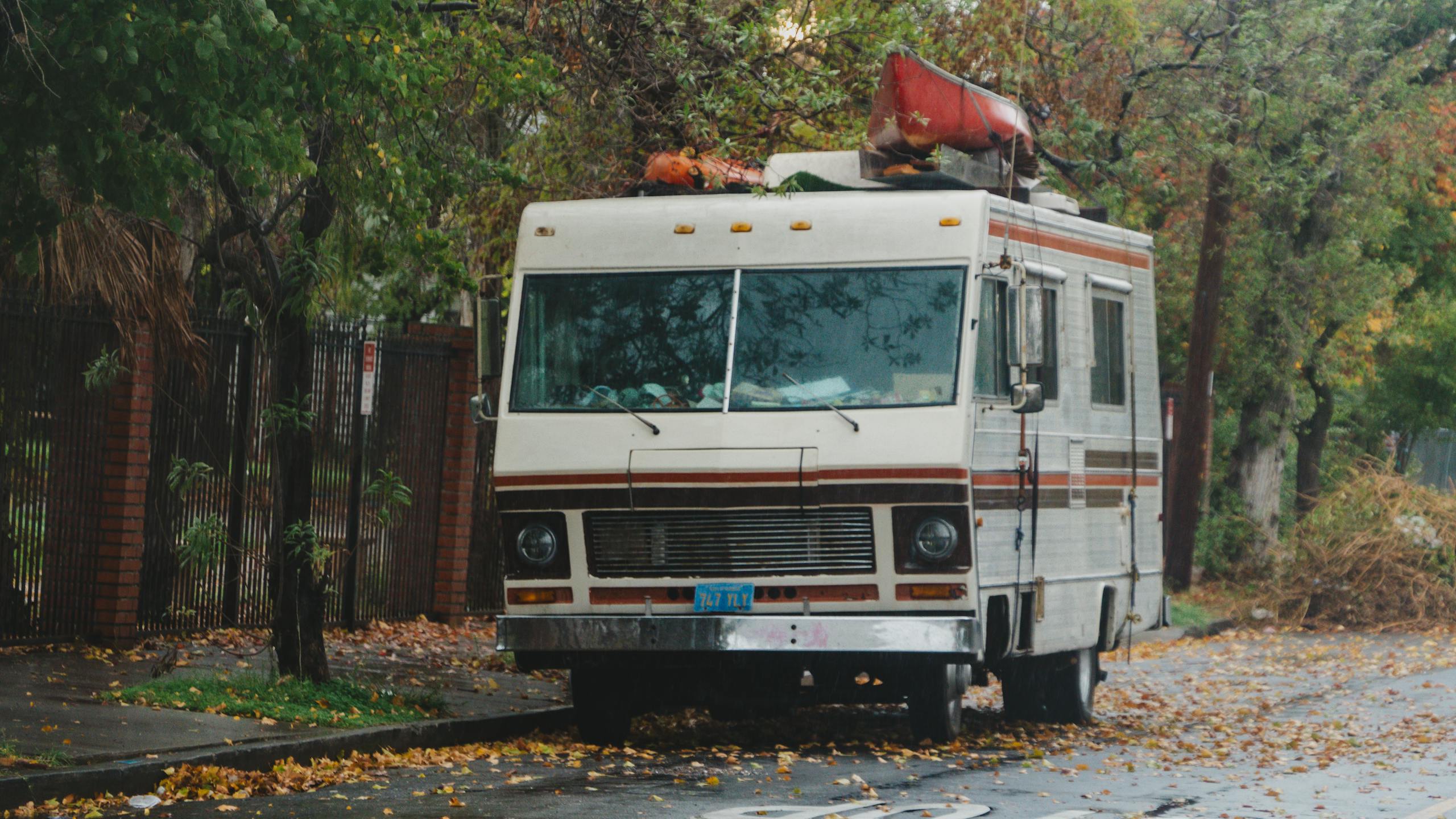 A vintage RV with a red canoe on top parked on a tree-lined street in an autumn setting.