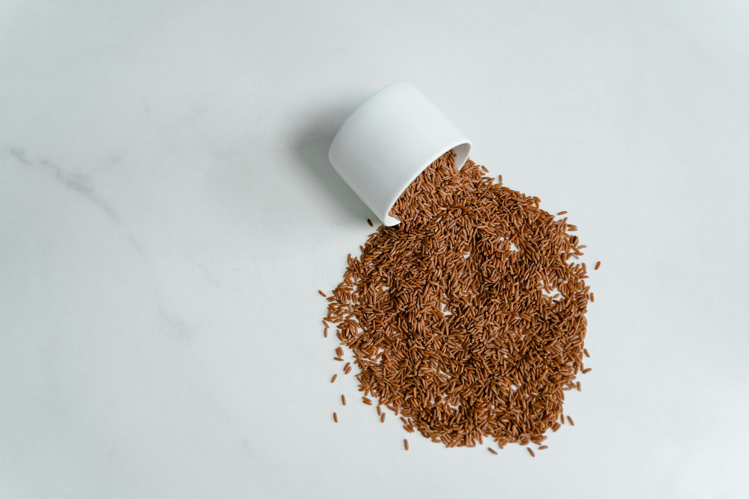 A white cup spills brown rice onto a kitchen countertop, showcasing a healthy food concept.