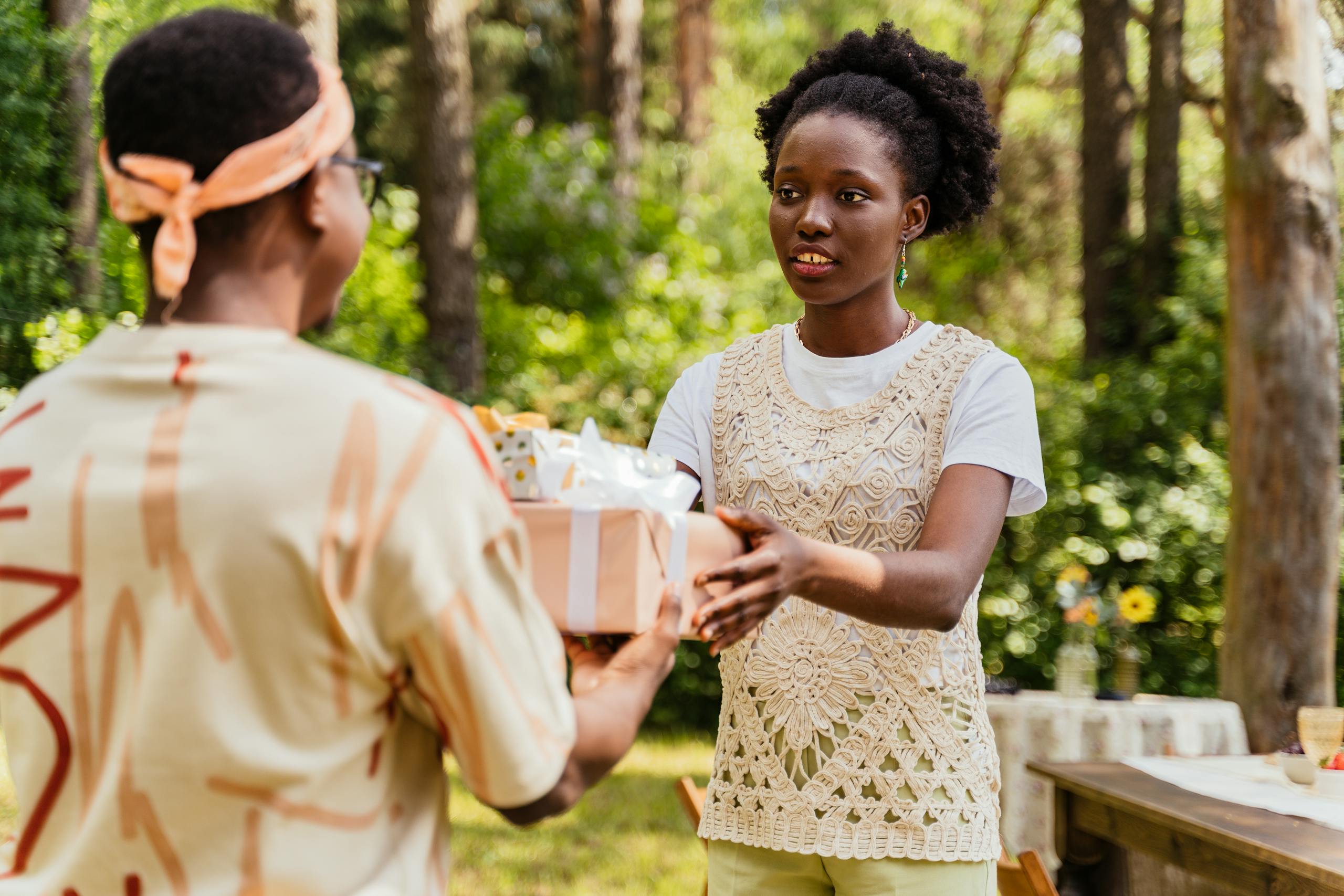 A woman hands a gift to another person during an outdoor celebration in a forest setting.