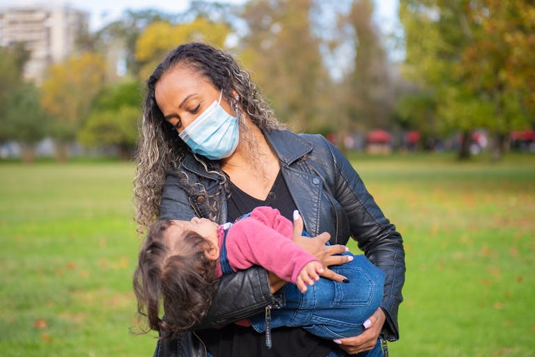 A woman in a leather jacket holds her child in a park while wearing a face mask.