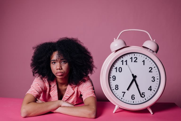 A woman in a pink shirt leans on a table beside a large pink alarm clock, set against a pink background.