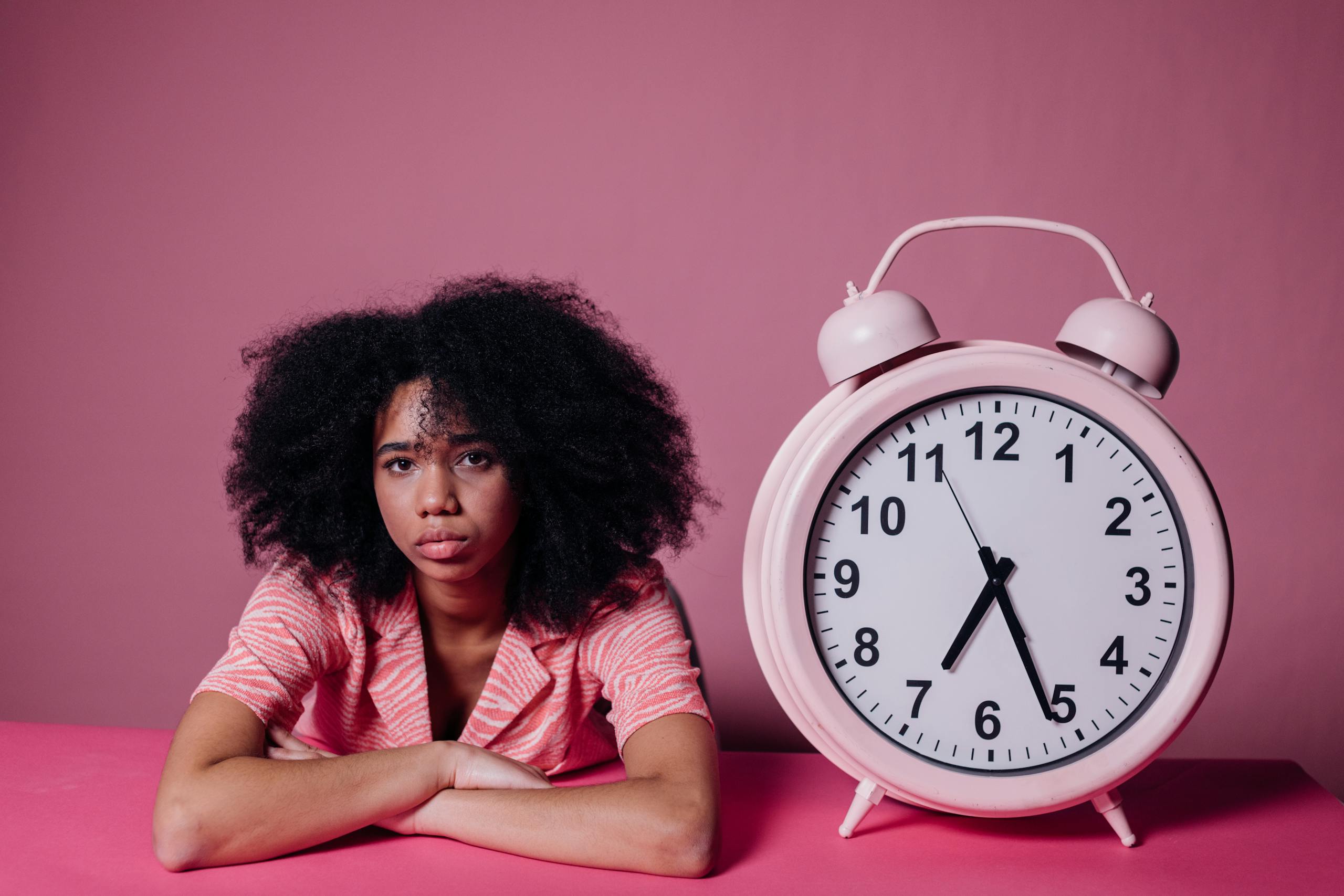 A woman in a pink shirt leans on a table beside a large pink alarm clock, set against a pink background.