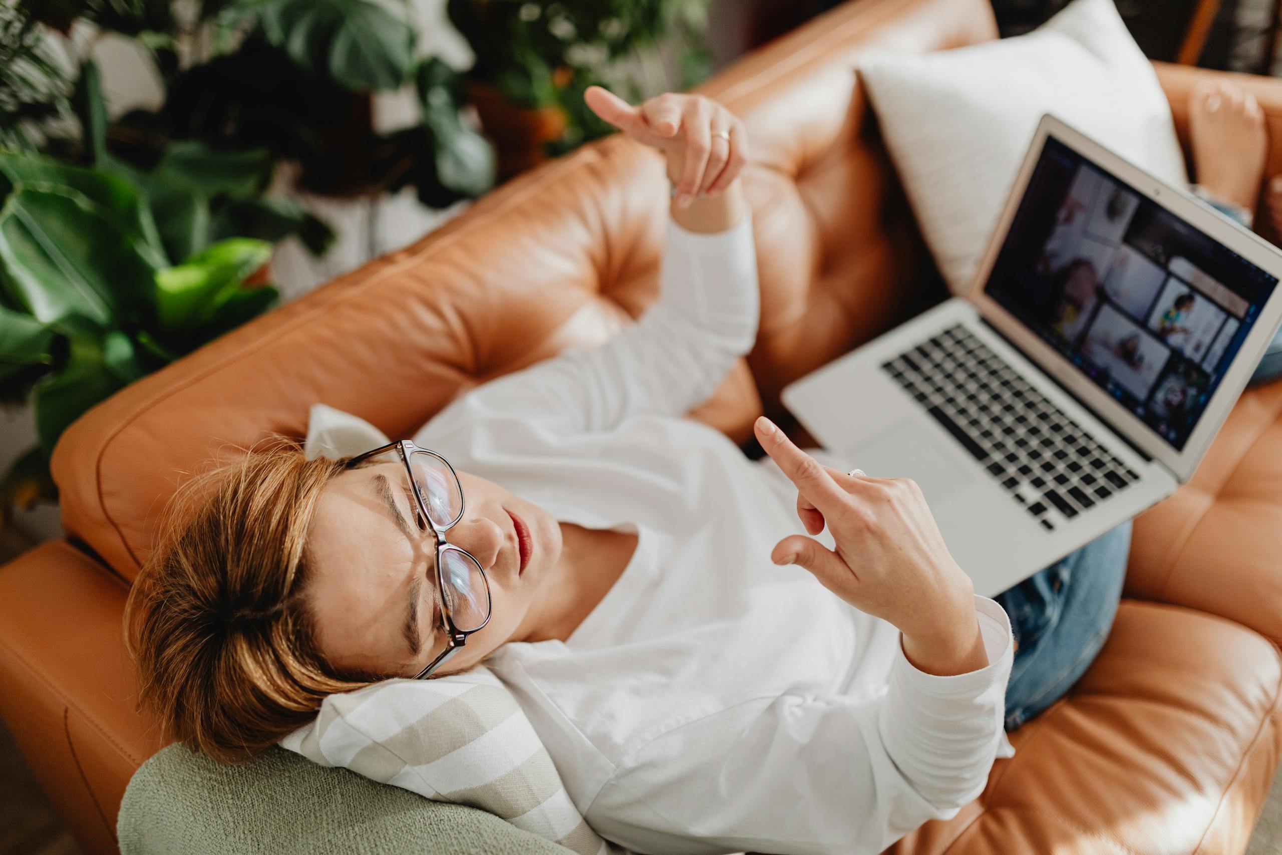 A woman in a white shirt lounges on a sofa with a laptop, surrounded by plants, enjoying leisure time.