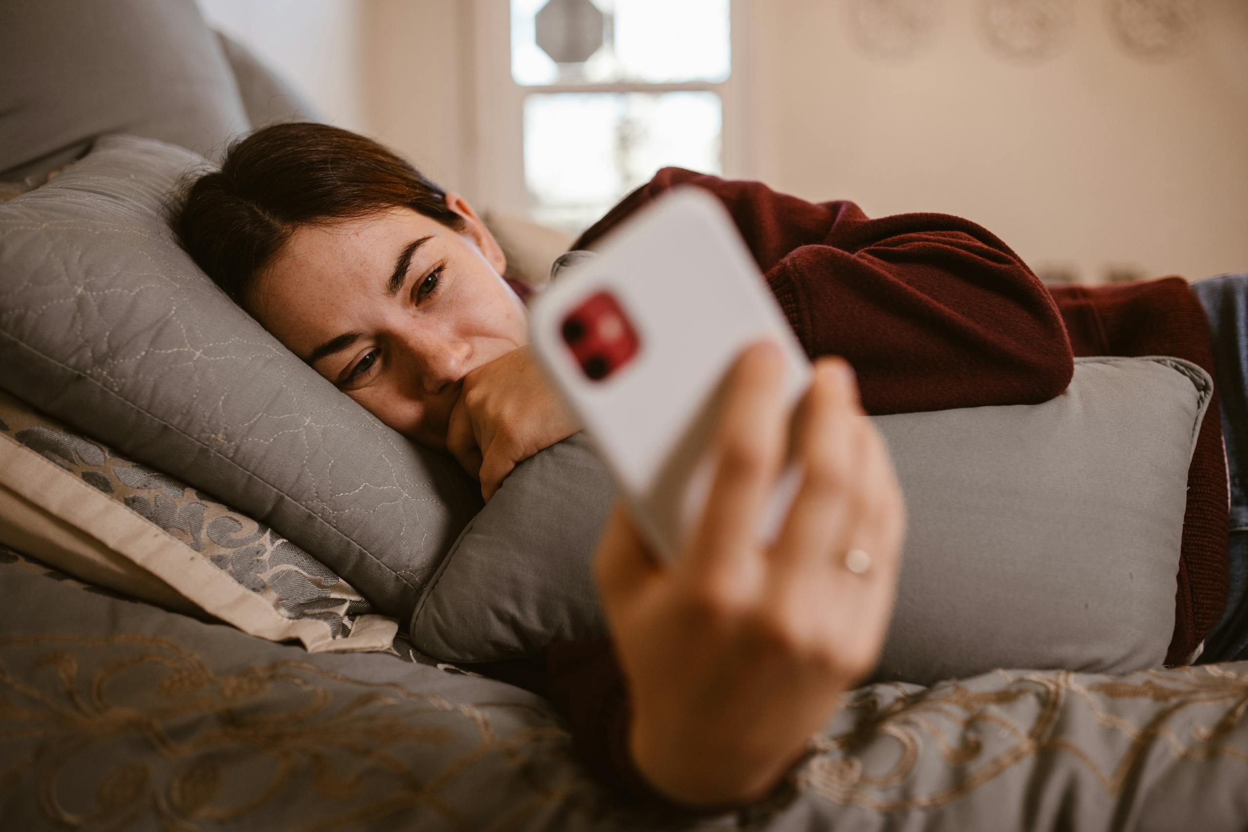 A woman lies comfortably on a bed, using her smartphone in a cozy indoor setting.