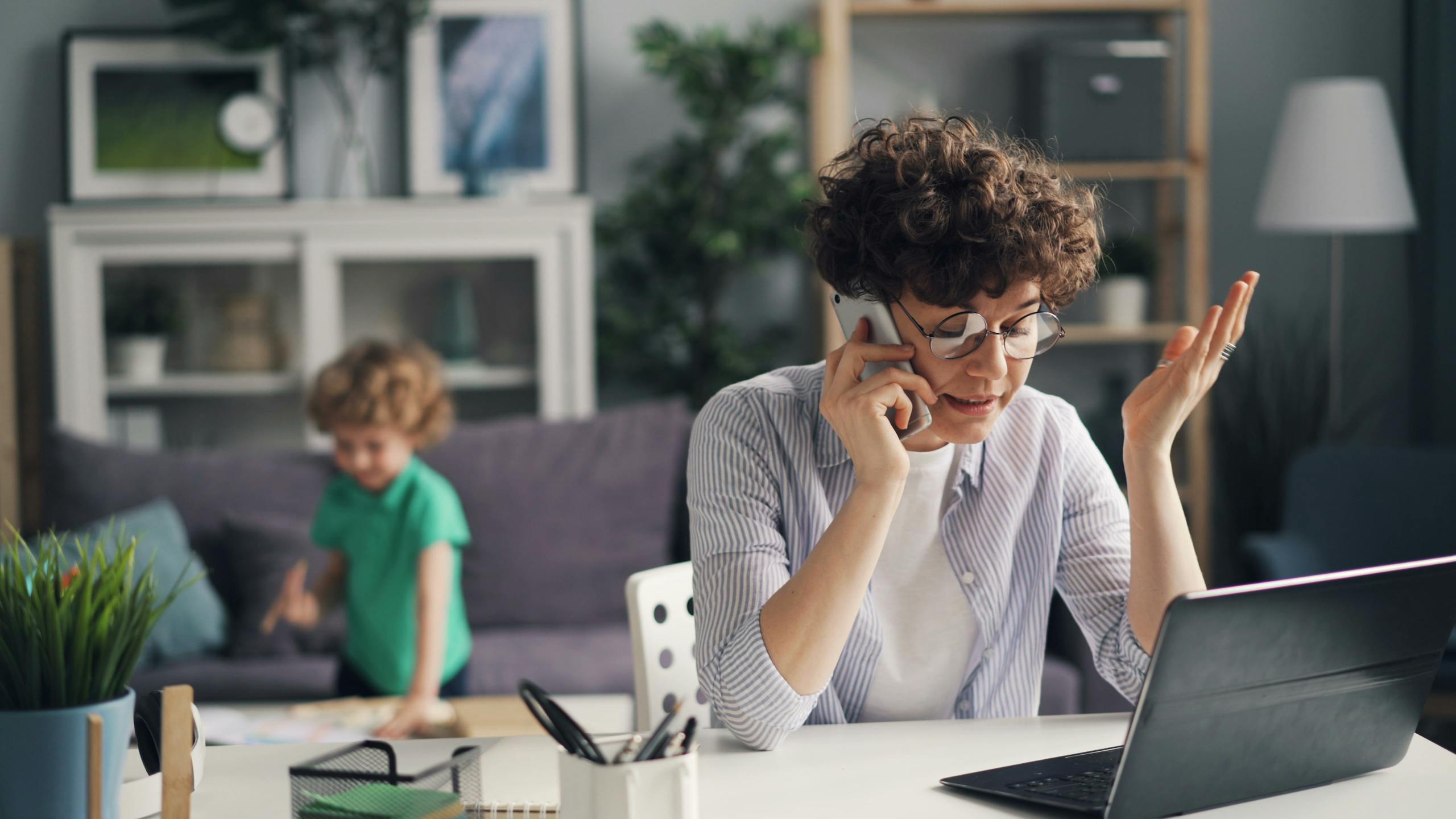 A woman multitasking with a phone and laptop while her child plays in the background.
