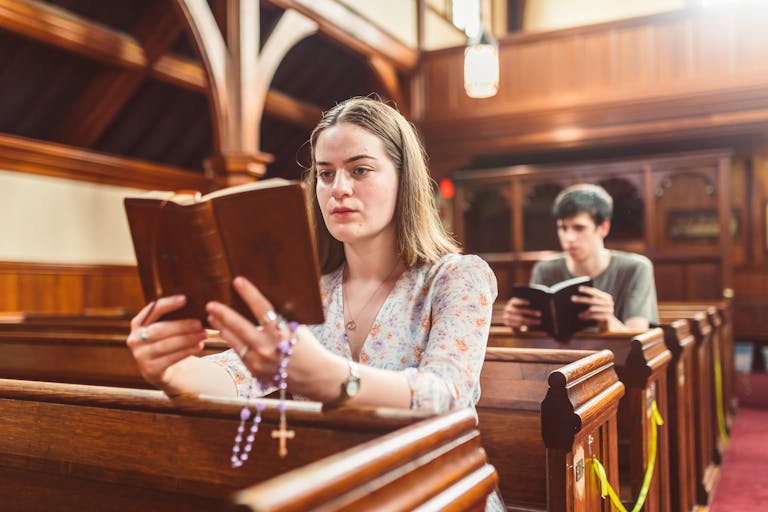 A woman reads a Bible and prays with rosary beads in a church pew.