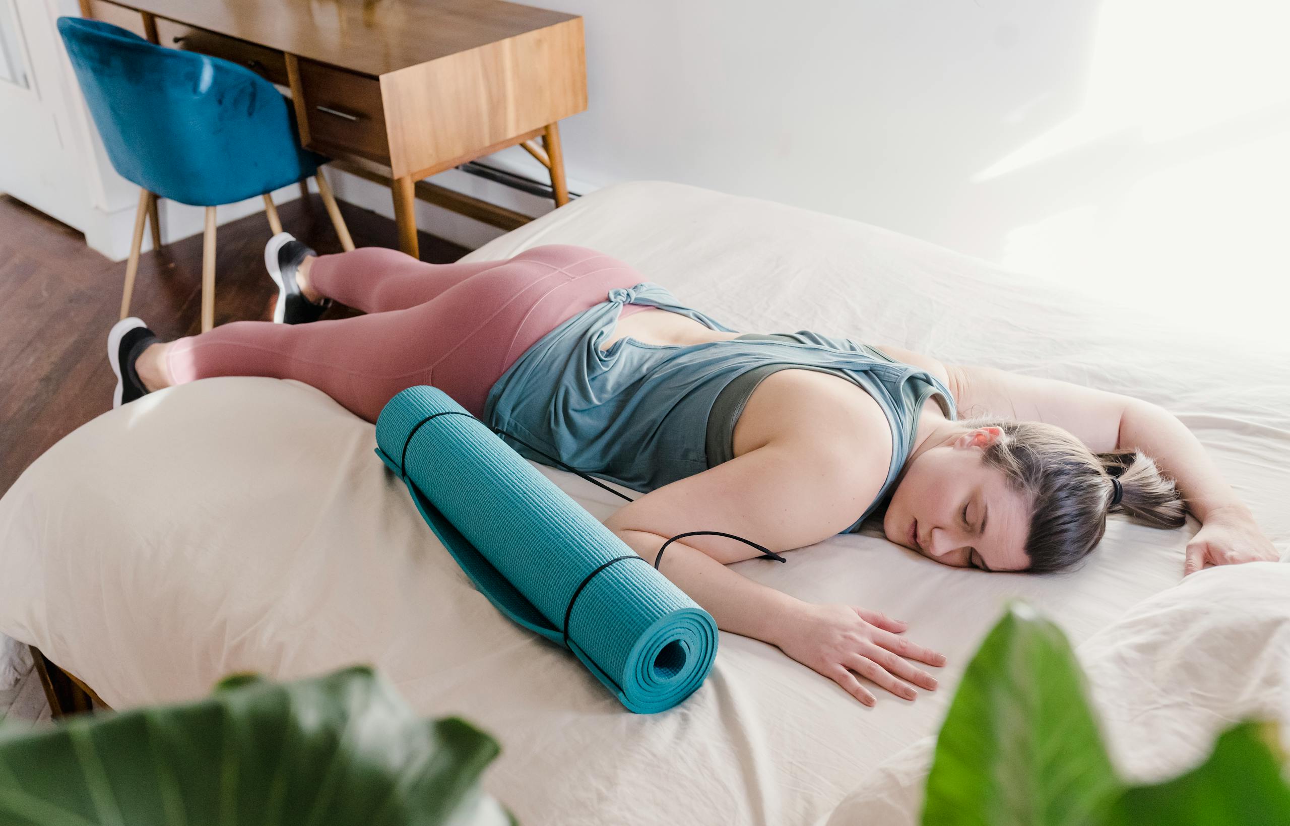 A woman rests on a bed with a yoga mat next to her, embracing relaxation and comfort indoors.