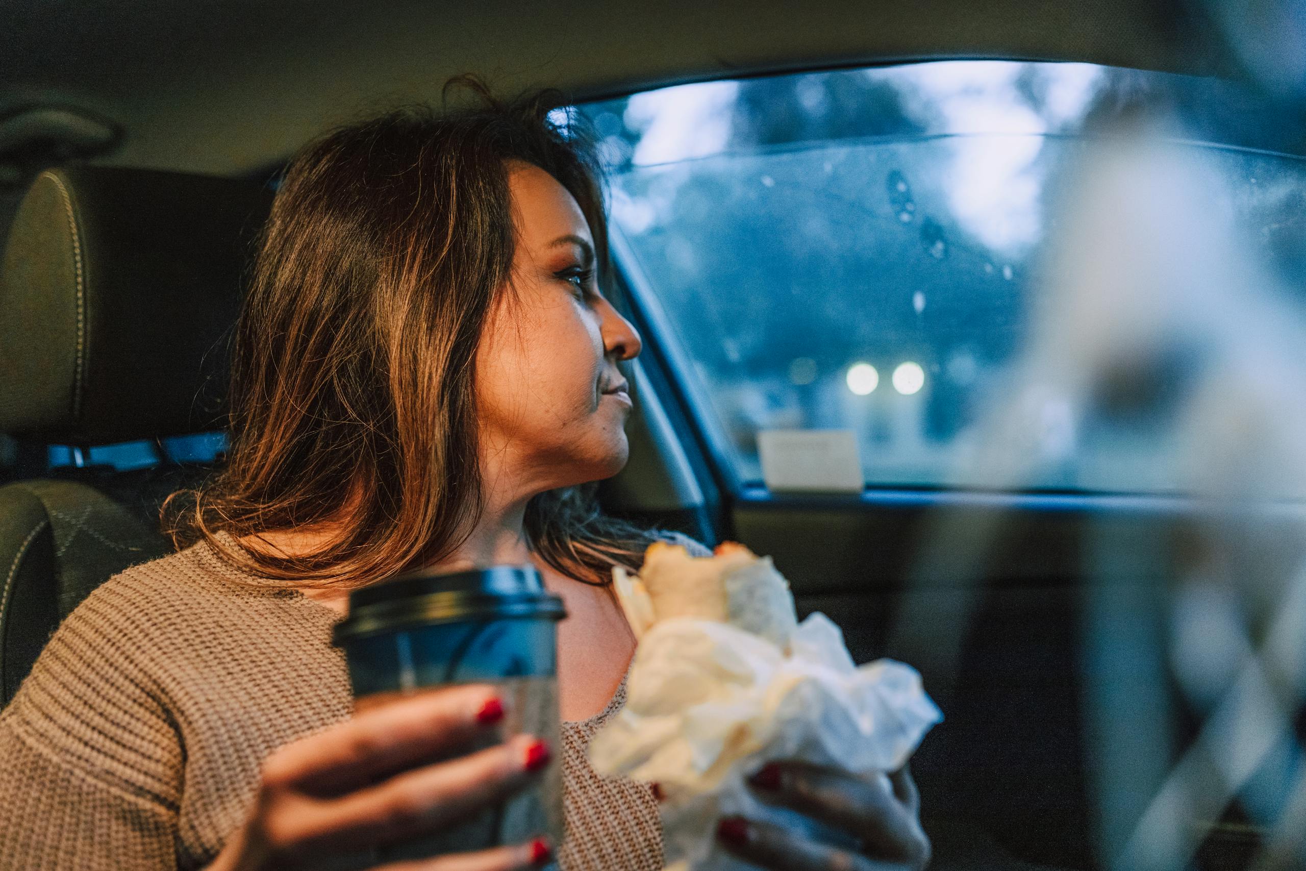 A woman sits in her car with a coffee and a sandwich, reflecting on her day.