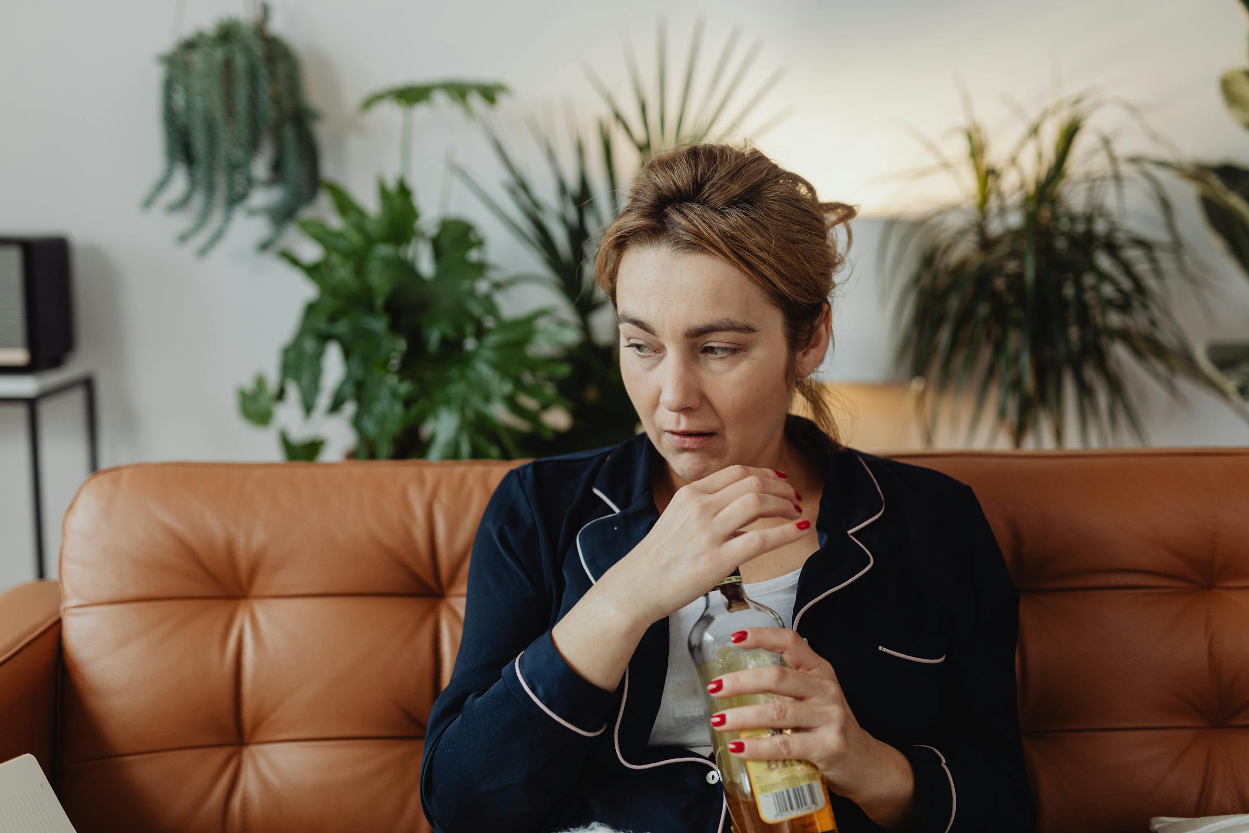 A woman sits pensively on a sofa with a bottle, surrounded by indoor plants.