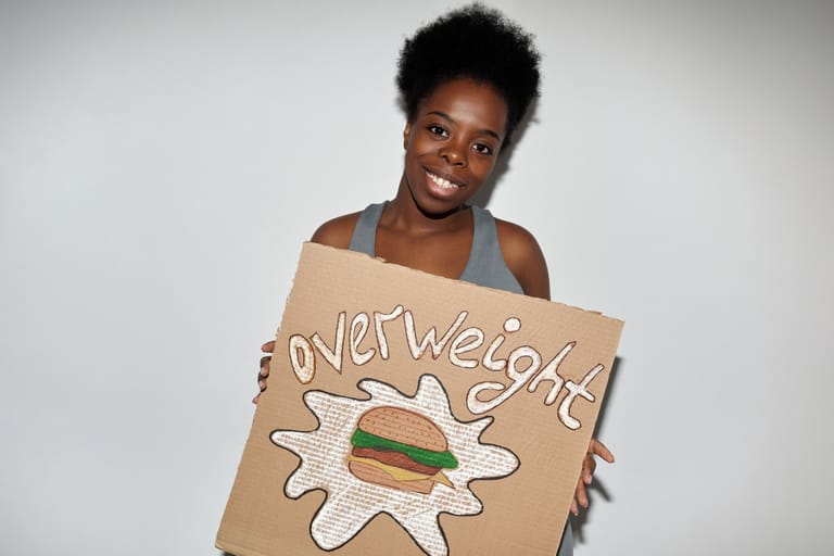 A woman smiles while holding a handmade cardboard sign with an artistic hamburger and the word overweight.