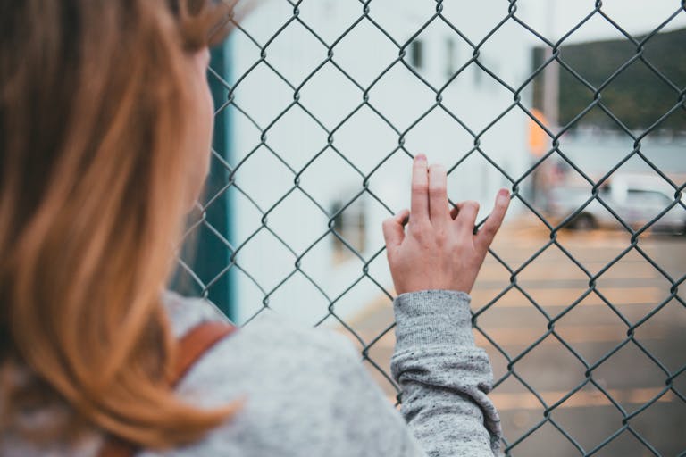 A woman stands outdoors, touching a chain-link fence with defocused background.
