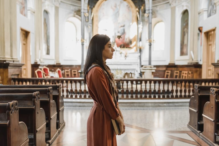 A woman stands solemnly in a beautifully lit, historic church lined with pews.