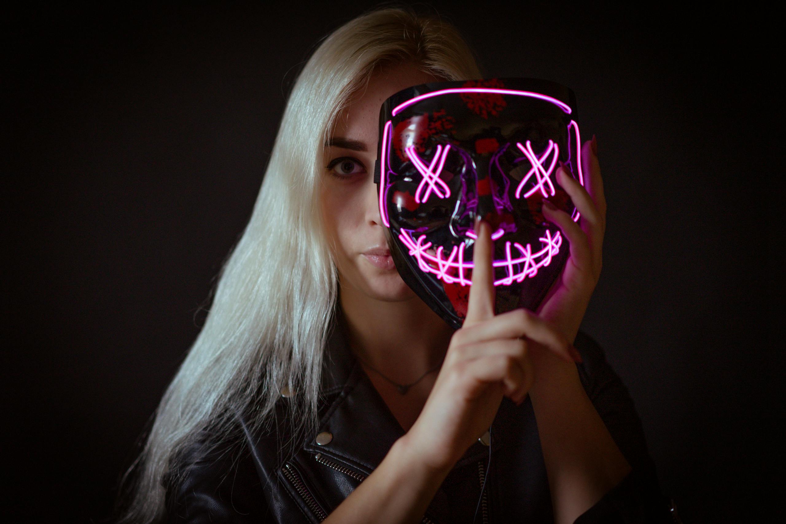A woman with blonde hair holds an illuminated neon mask, conveying mystery and secrecy.