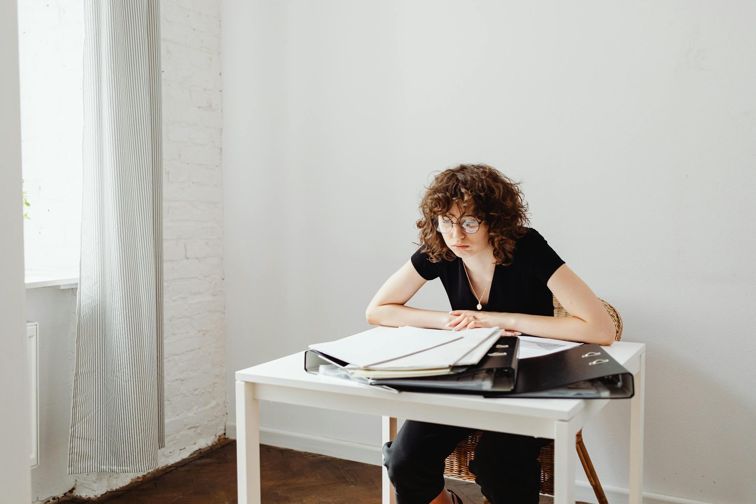 A woman with curly hair reads documents at a white desk indoors.
