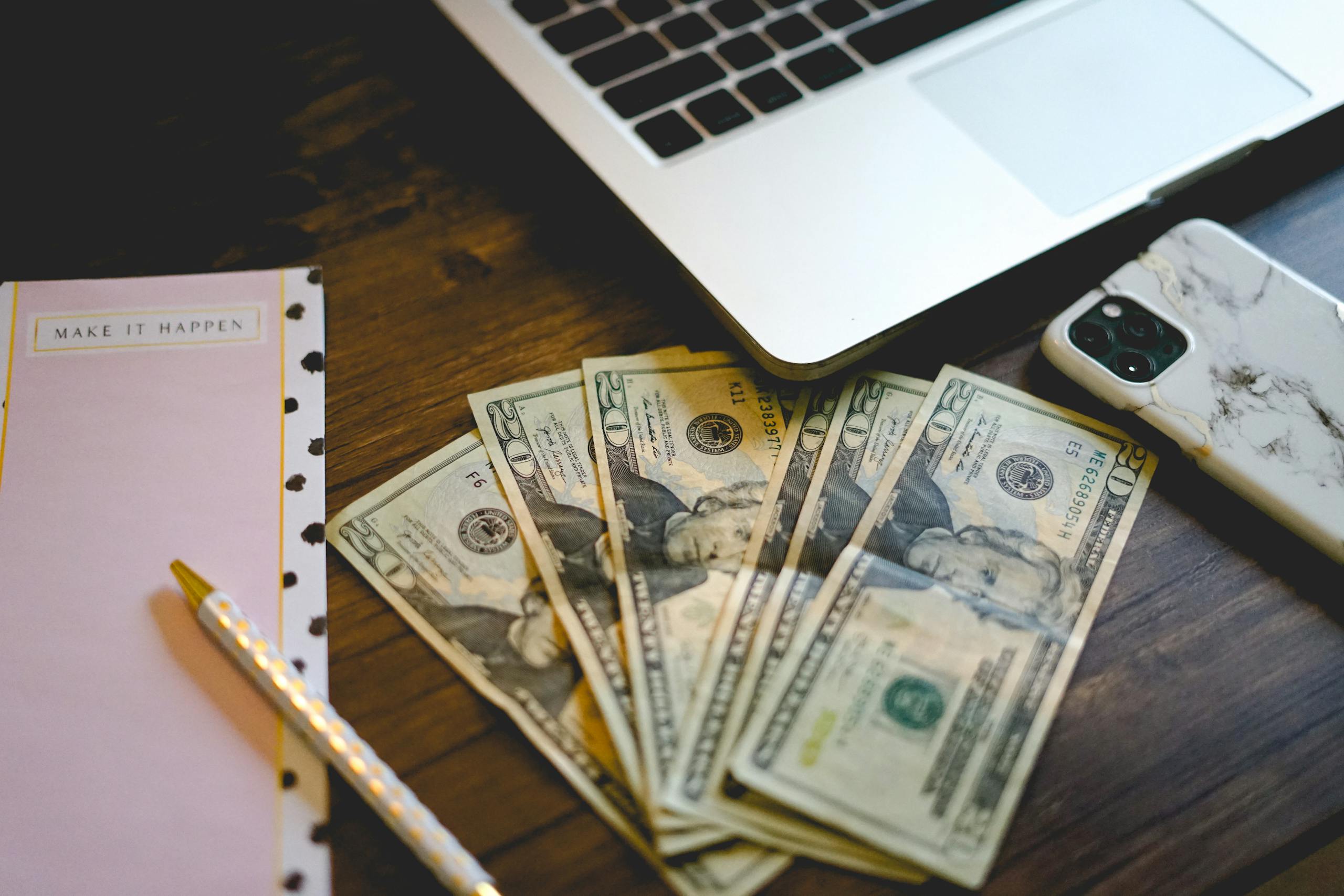 A workspace setup with dollar bills, notepad, and laptop on a wooden desk.