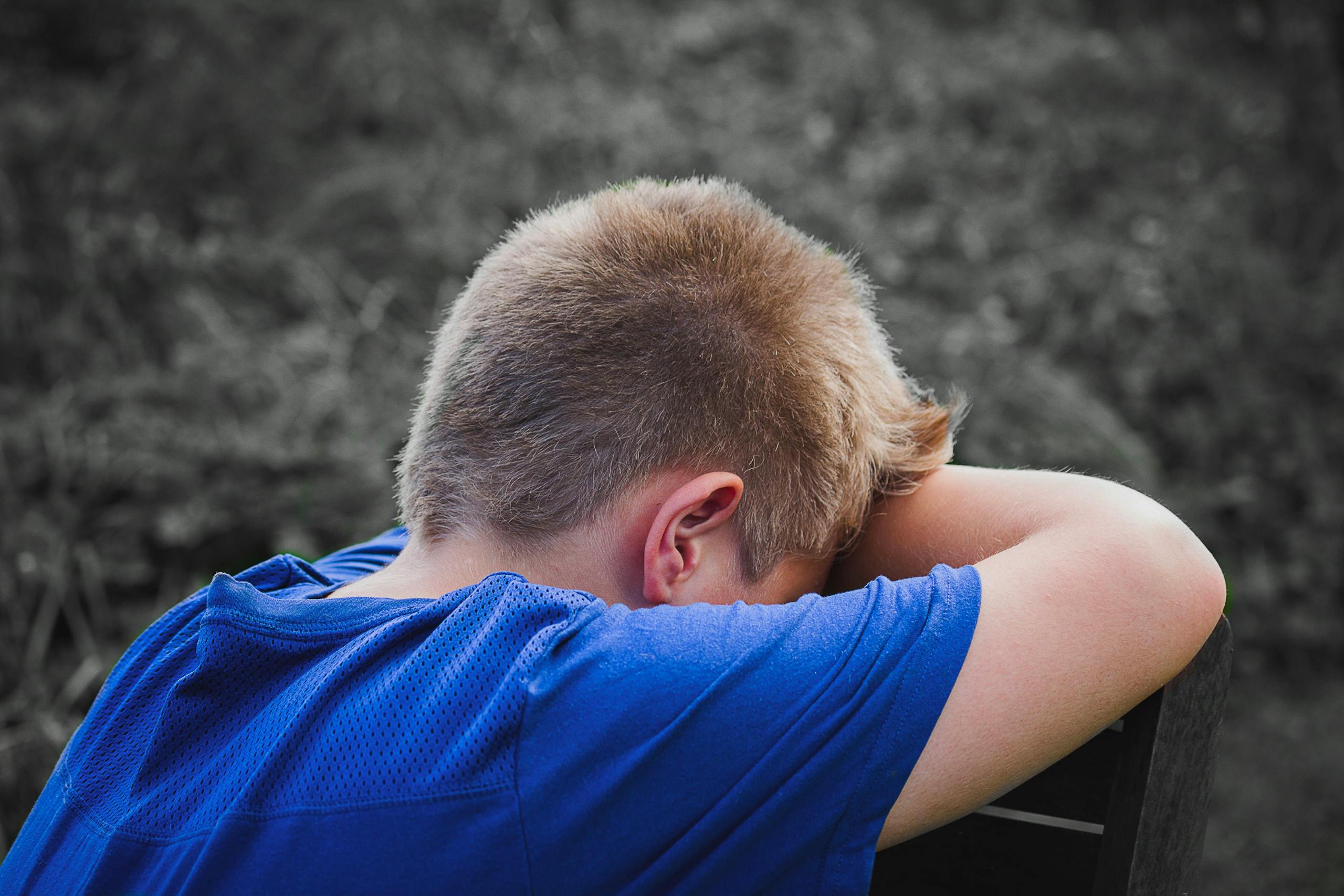 A young boy in a blue shirt rests his head on his arms outdoors, appearing sad and contemplative.