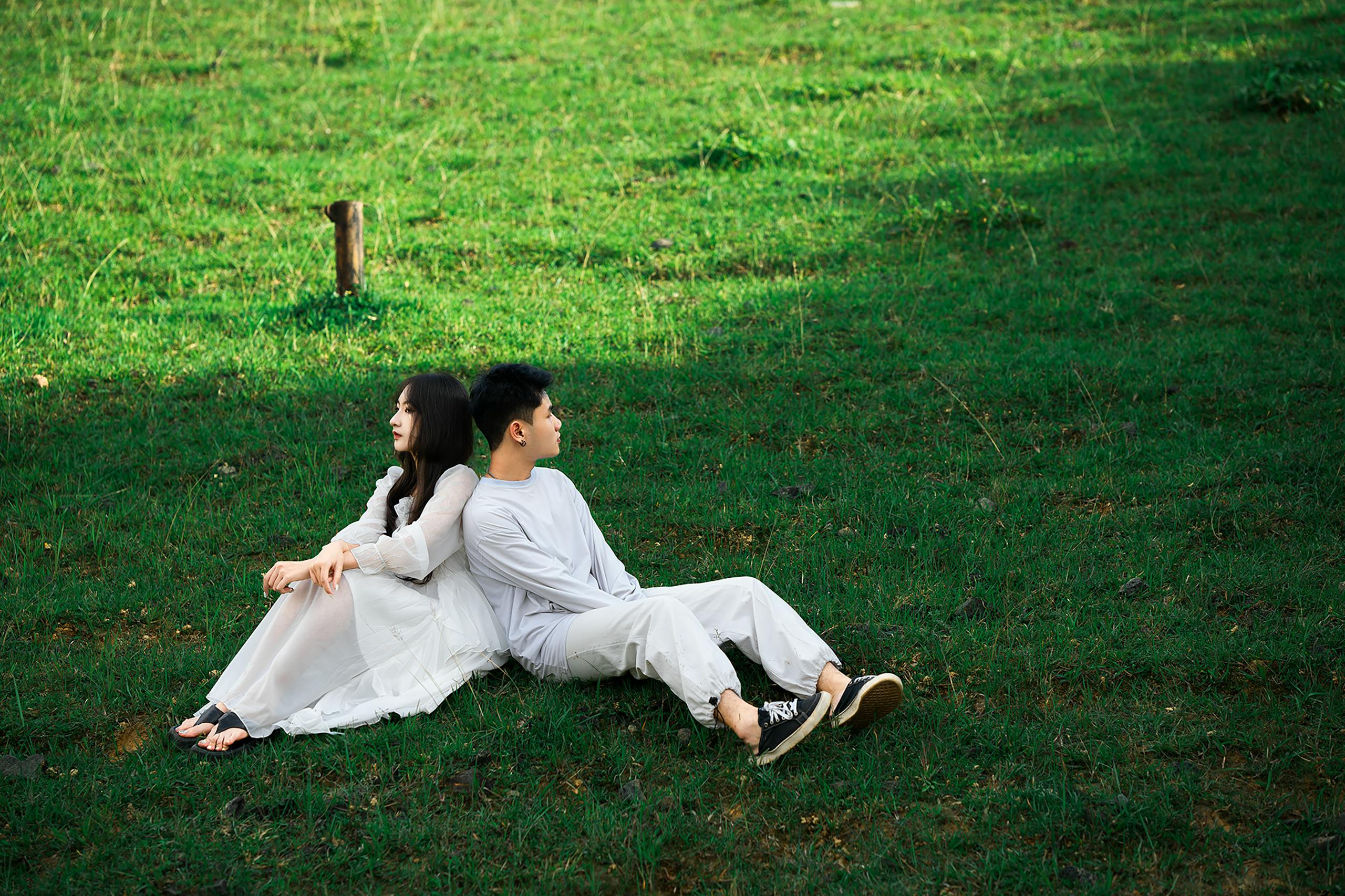 A young couple sits back to back on lush green grass in Dalat, Vietnam, enjoying leisure time outdoors.