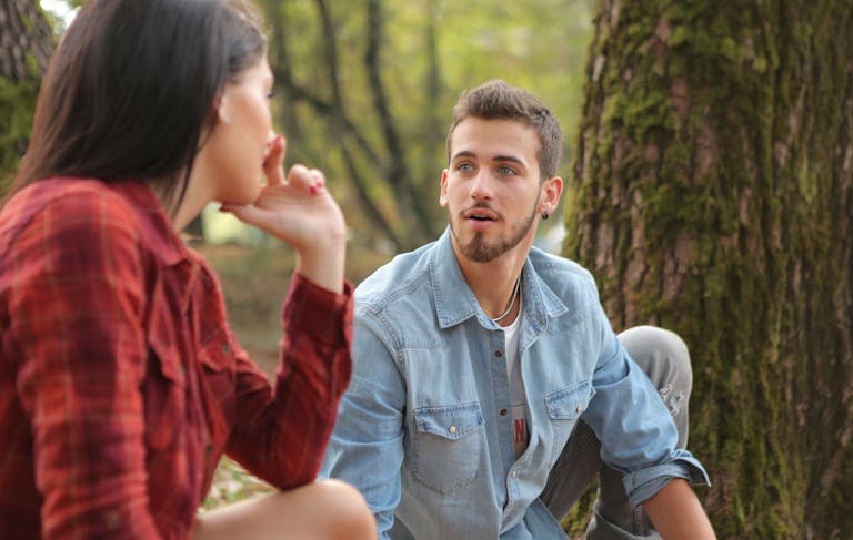 A young couple sitting and talking in a lush forest, showcasing casual fashion and natural environment.