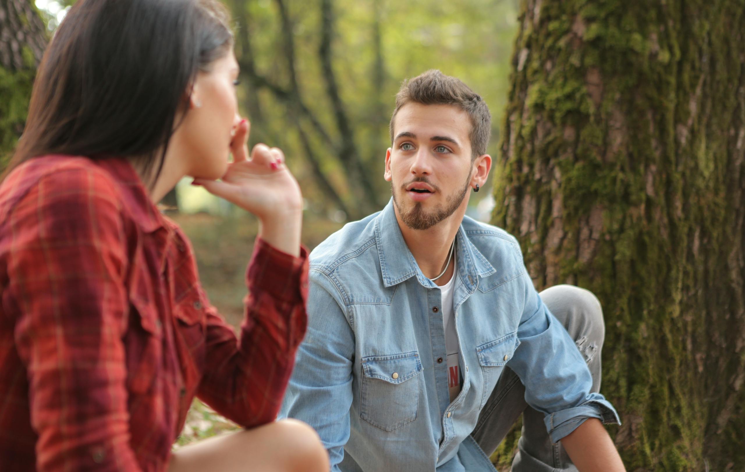 A young couple sitting and talking in a lush forest, showcasing casual fashion and natural environment.