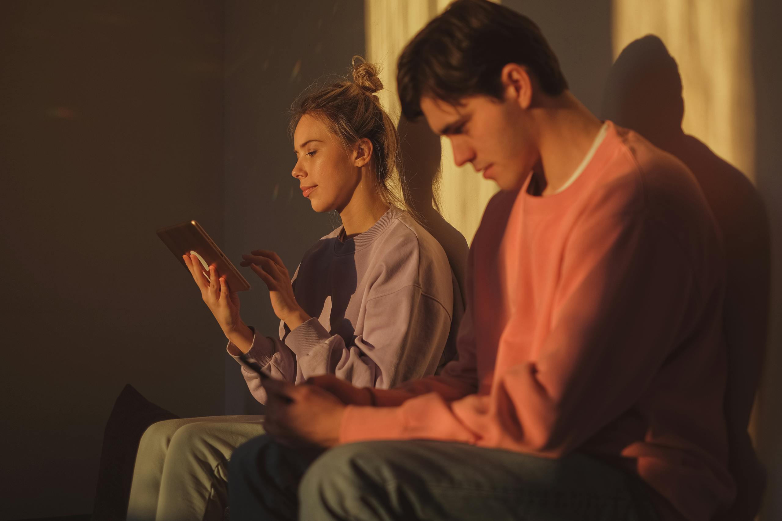 A young couple sitting together indoors, engaged with electronic devices in warm sunlight.