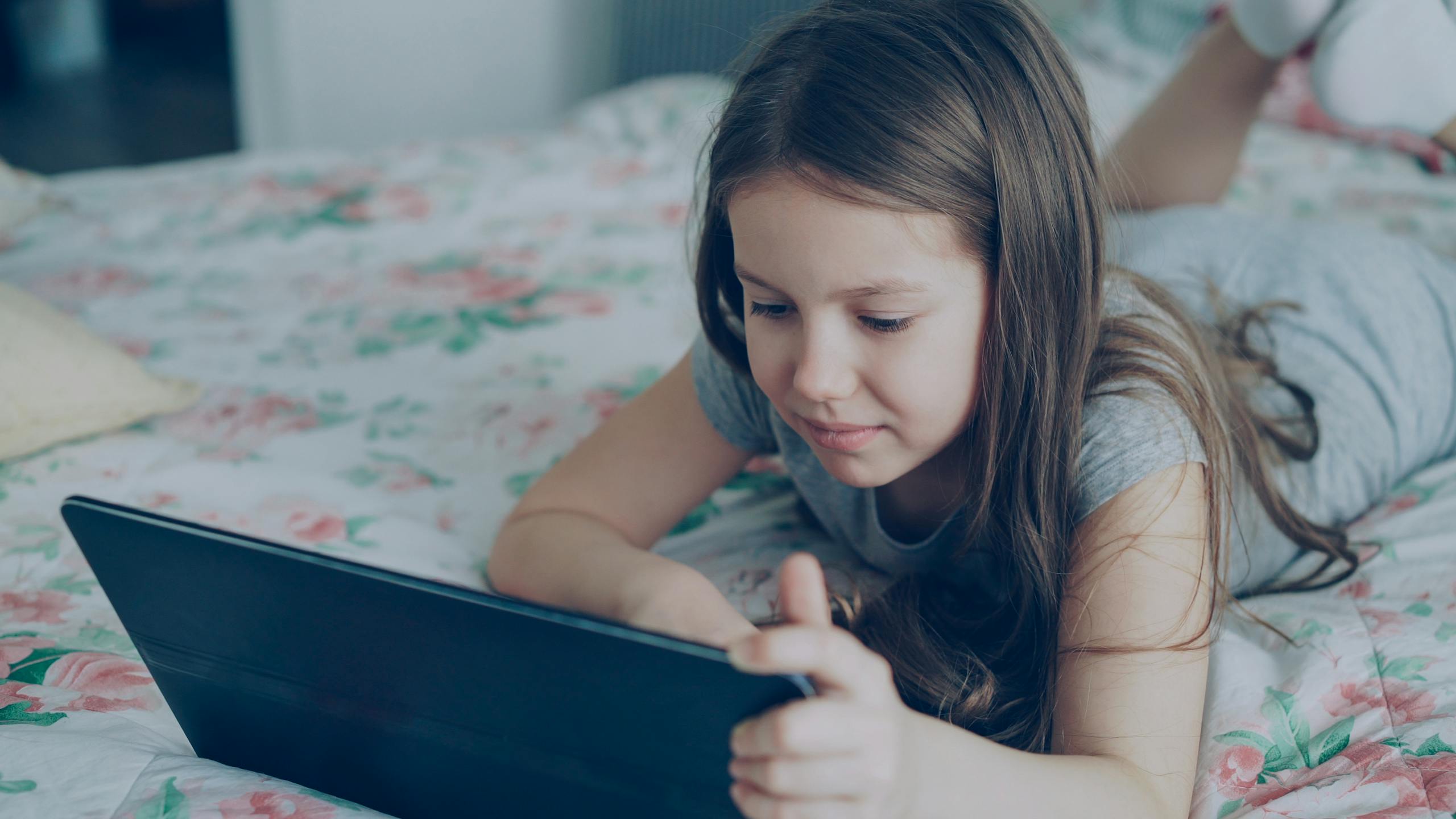 A young girl lies on a bed using a tablet, immersed in her digital world.