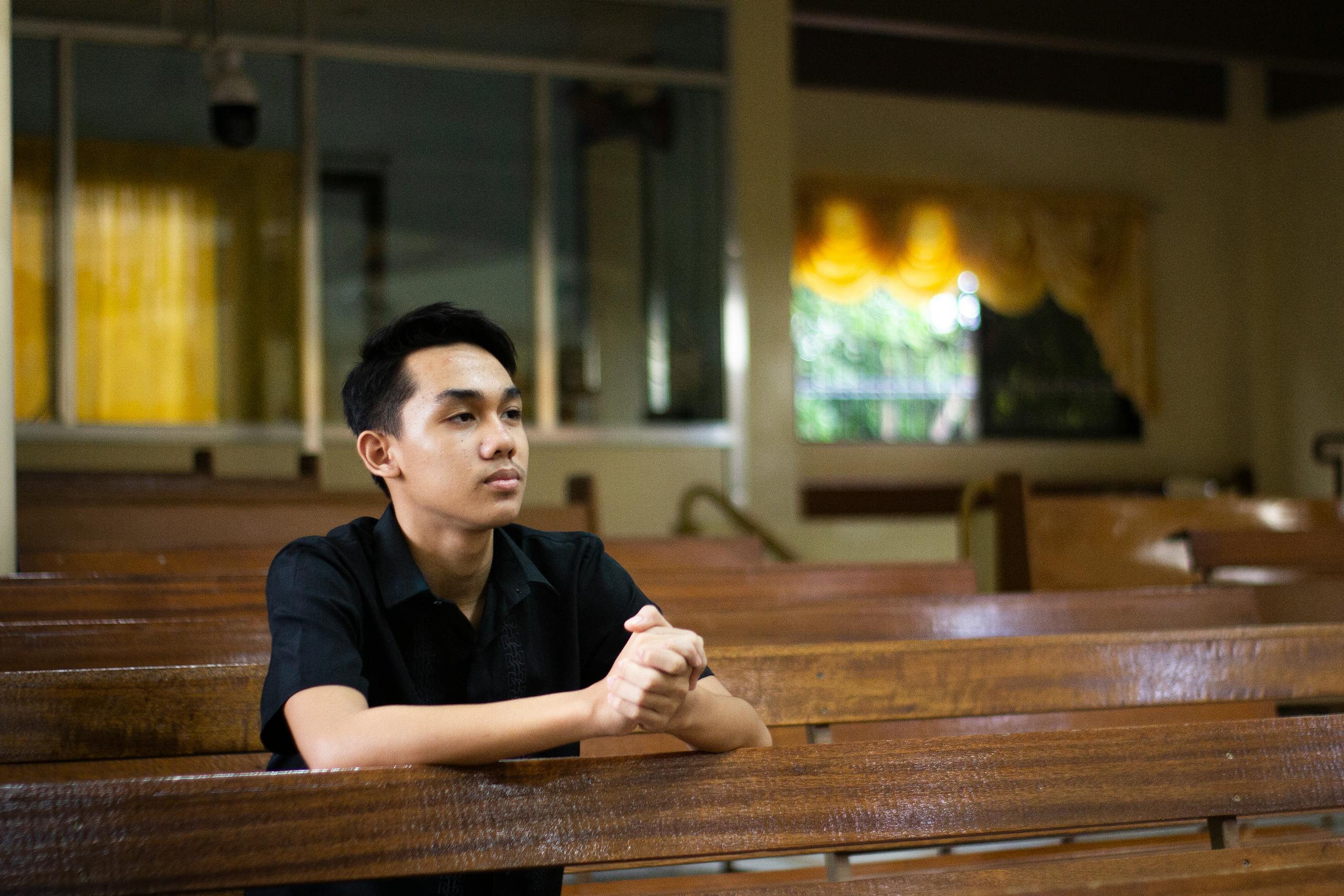 A young man in a church, sitting in a pew, clasping hands in prayer.