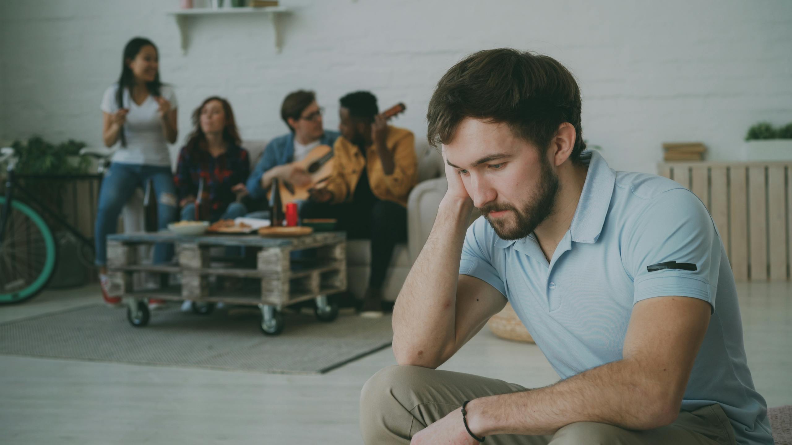 A young man sits alone looking somber while friends socialize in the background.