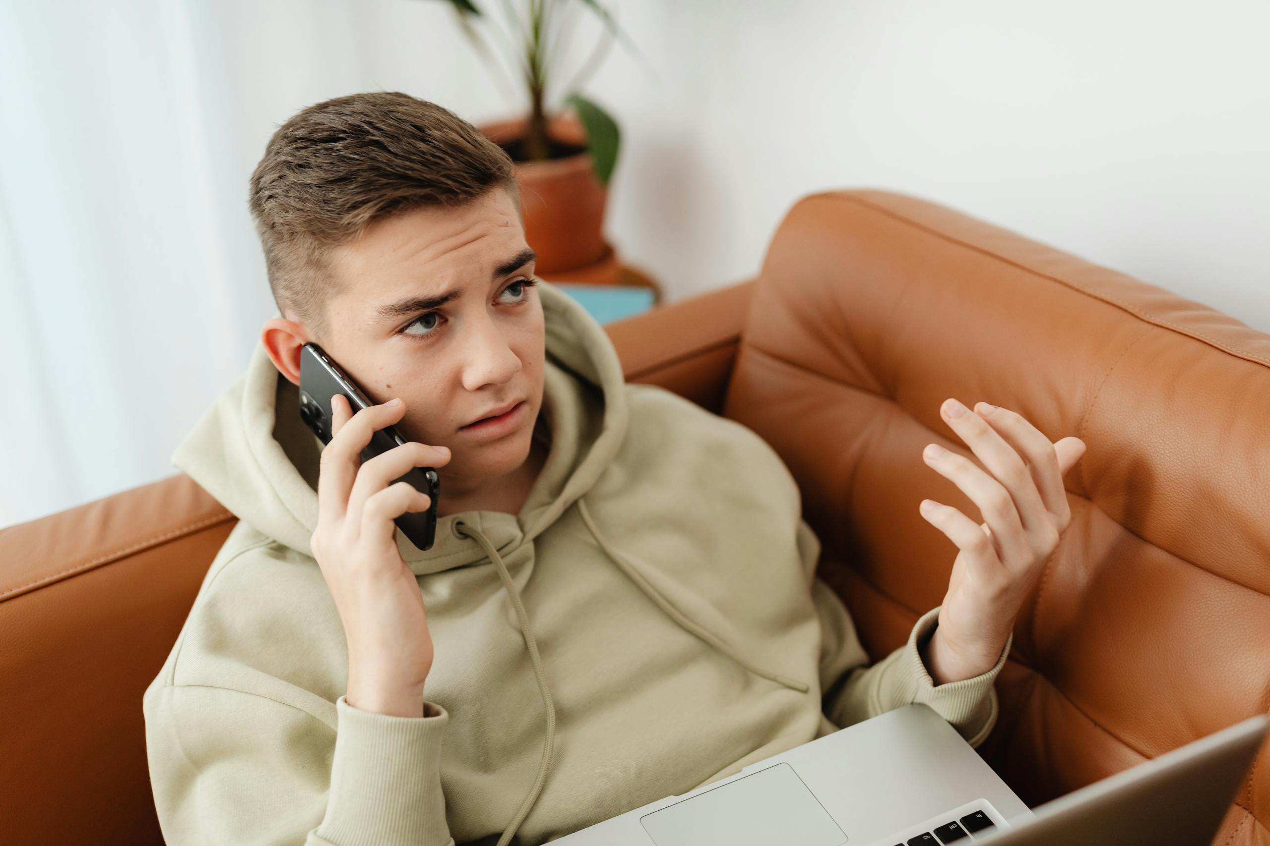 A young man talks on the phone while working on a laptop, looking concerned and frustrated.