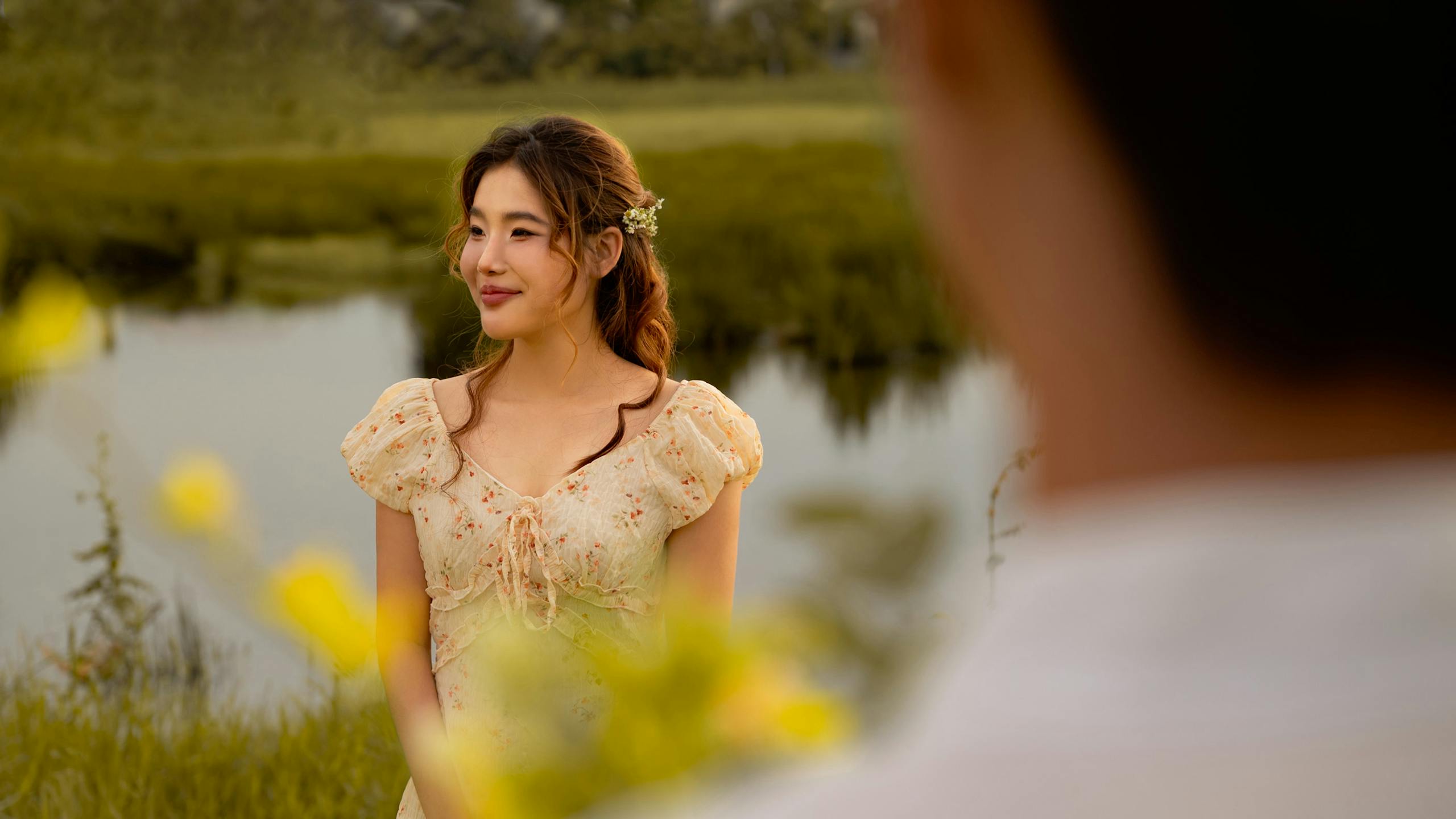 A young woman in a summer dress stands by a peaceful lake, exuding a sense of tranquility.