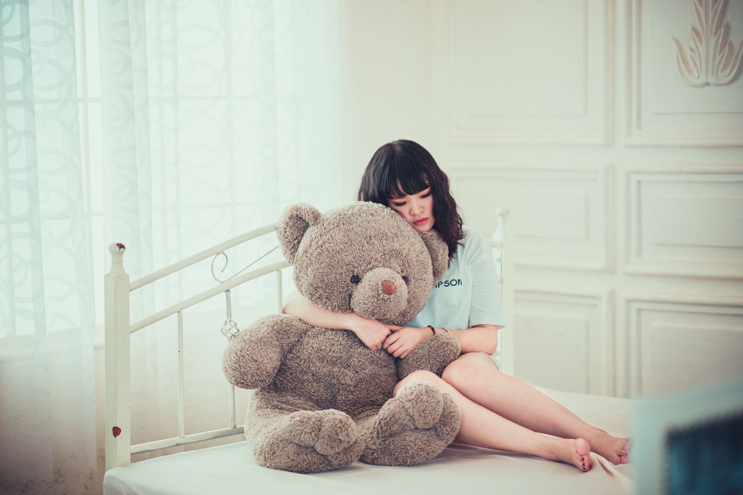 A young woman peacefully hugging a teddy bear while sitting on a bed in a sunlit room.