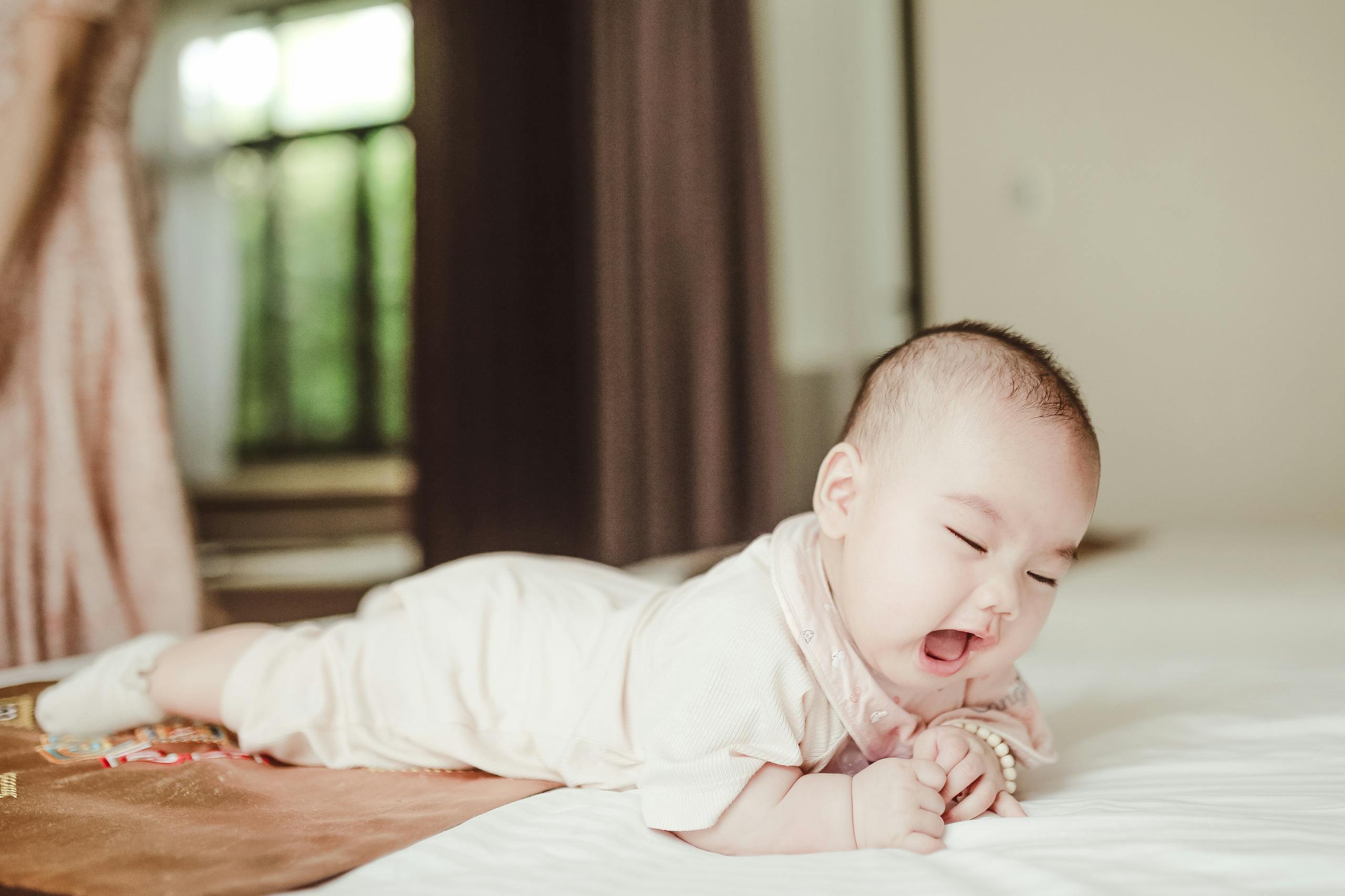 Adorable newborn baby in light pink bodysuit lying on belly on white soft coverlet on bed