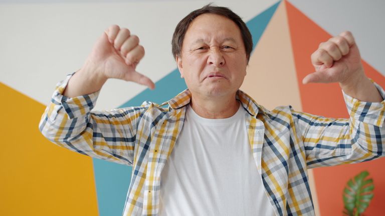 Adult male expressing disapproval with thumbs down gesture against a colorful geometric background indoors.
