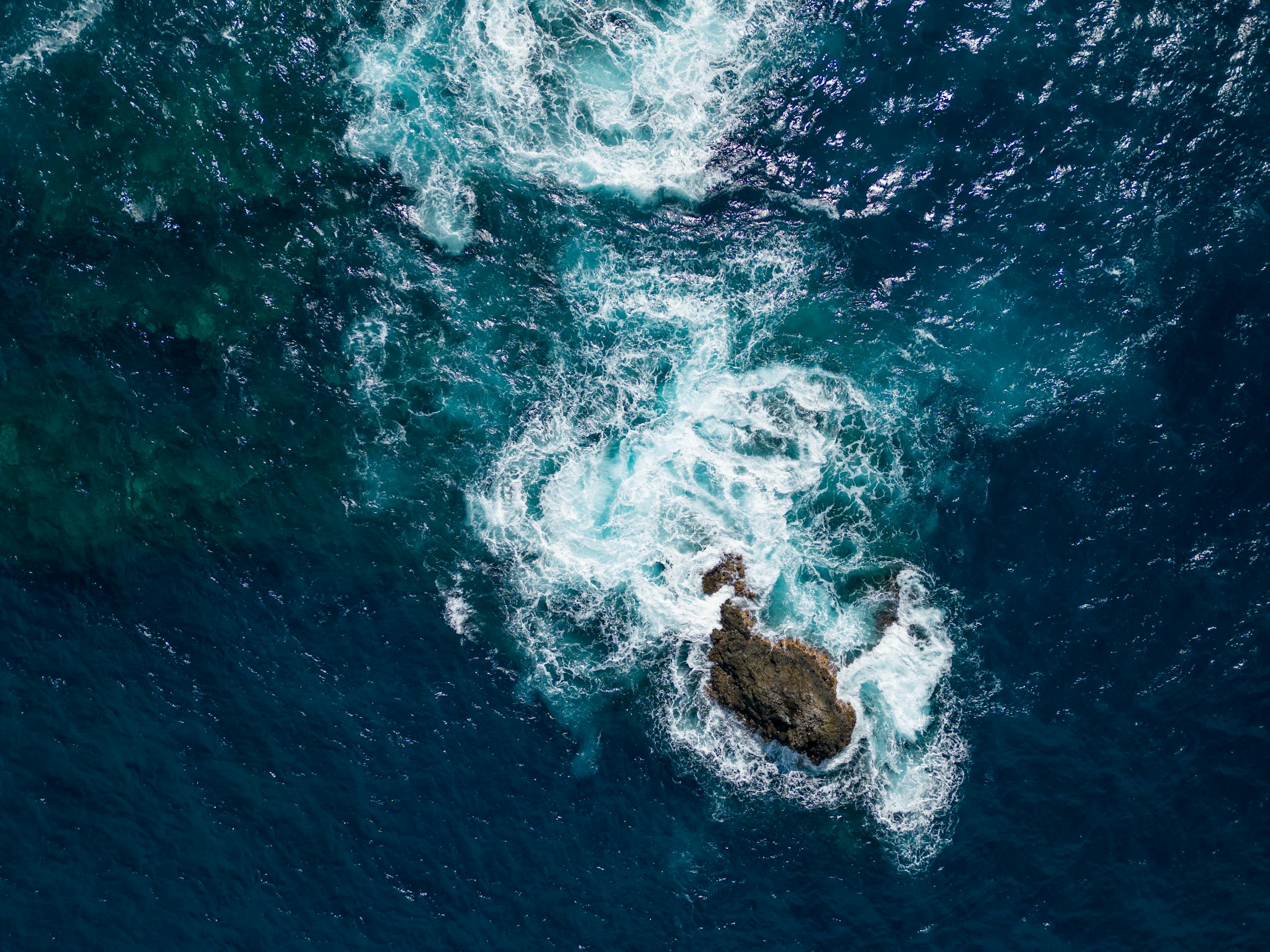 Aerial shot of powerful ocean waves crashing against a solitary rock formation, showcasing nature's raw beauty.