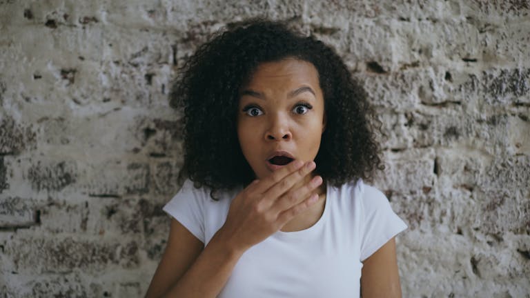 Amazed woman covering her mouth while standing by a rustic brick wall indoors.