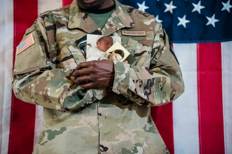 American soldier in uniform holding family photos against a flag backdrop.