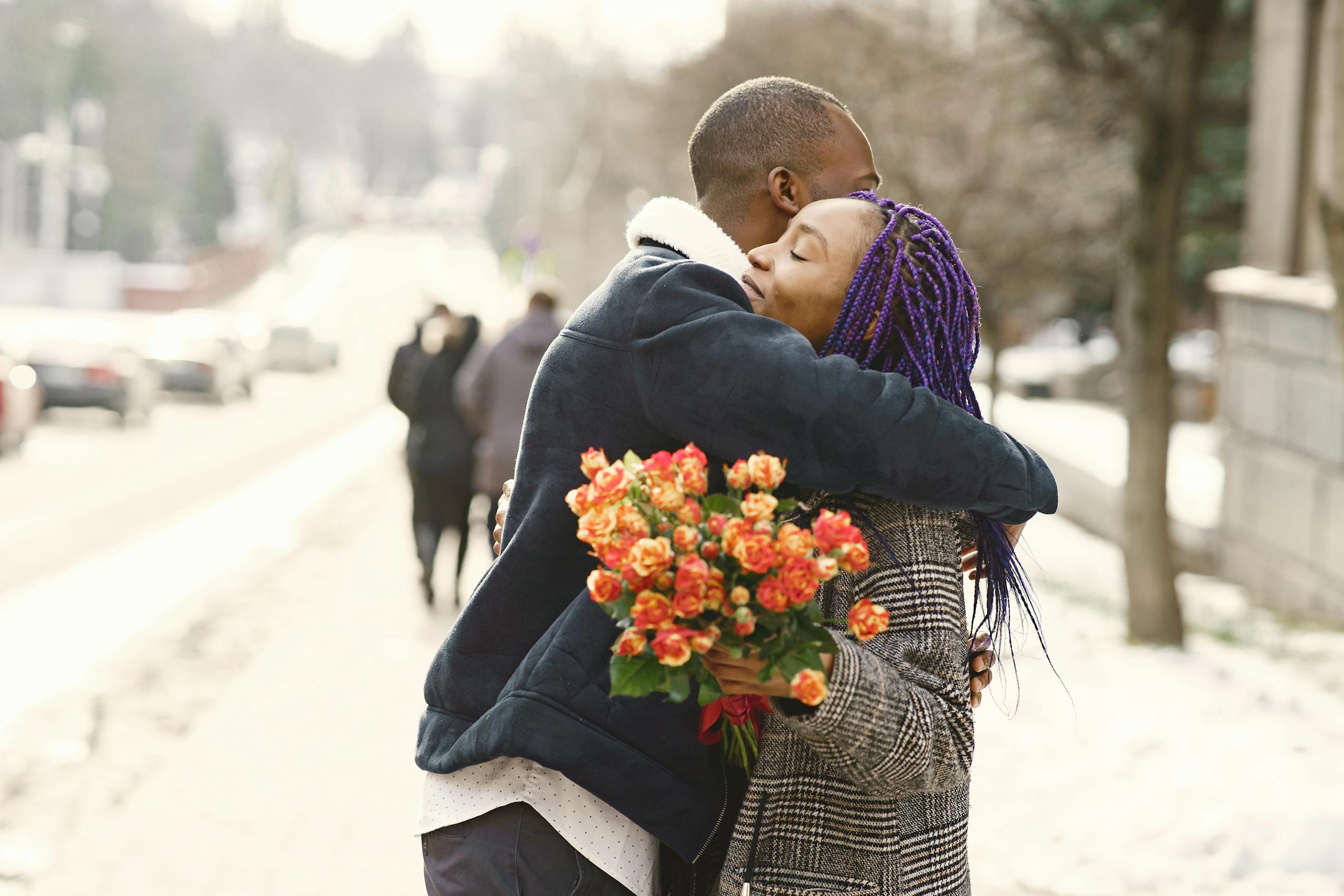 An affectionate couple shares a hug outdoors, holding a bouquet on a snowy street.