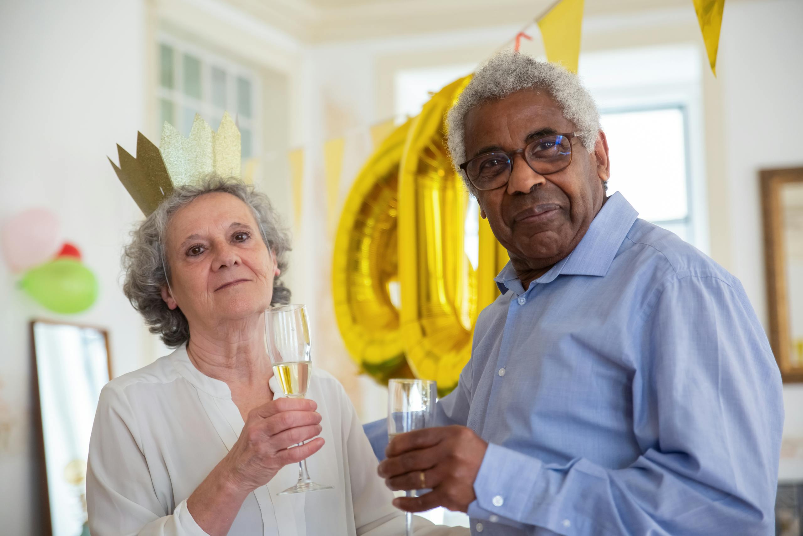 An elderly couple enjoying a festive celebration at home, holding champagne glasses with decorations in the background.