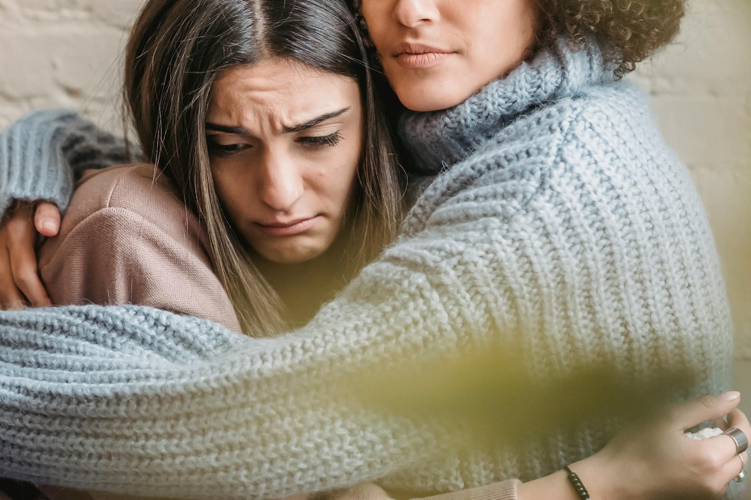 Anonymous woman in warn sweater comforting and embracing disappointed female friend near wall in room at home with green leaf