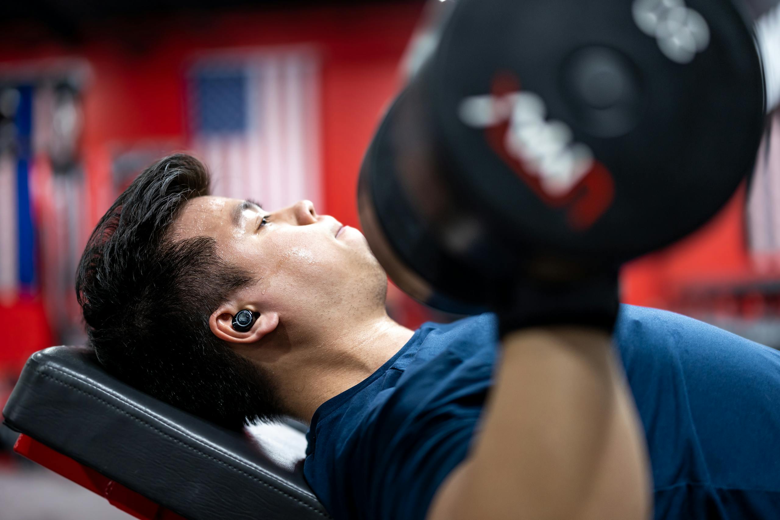Asian man lifting dumbbells on a bench in a gym, focusing on strength training.