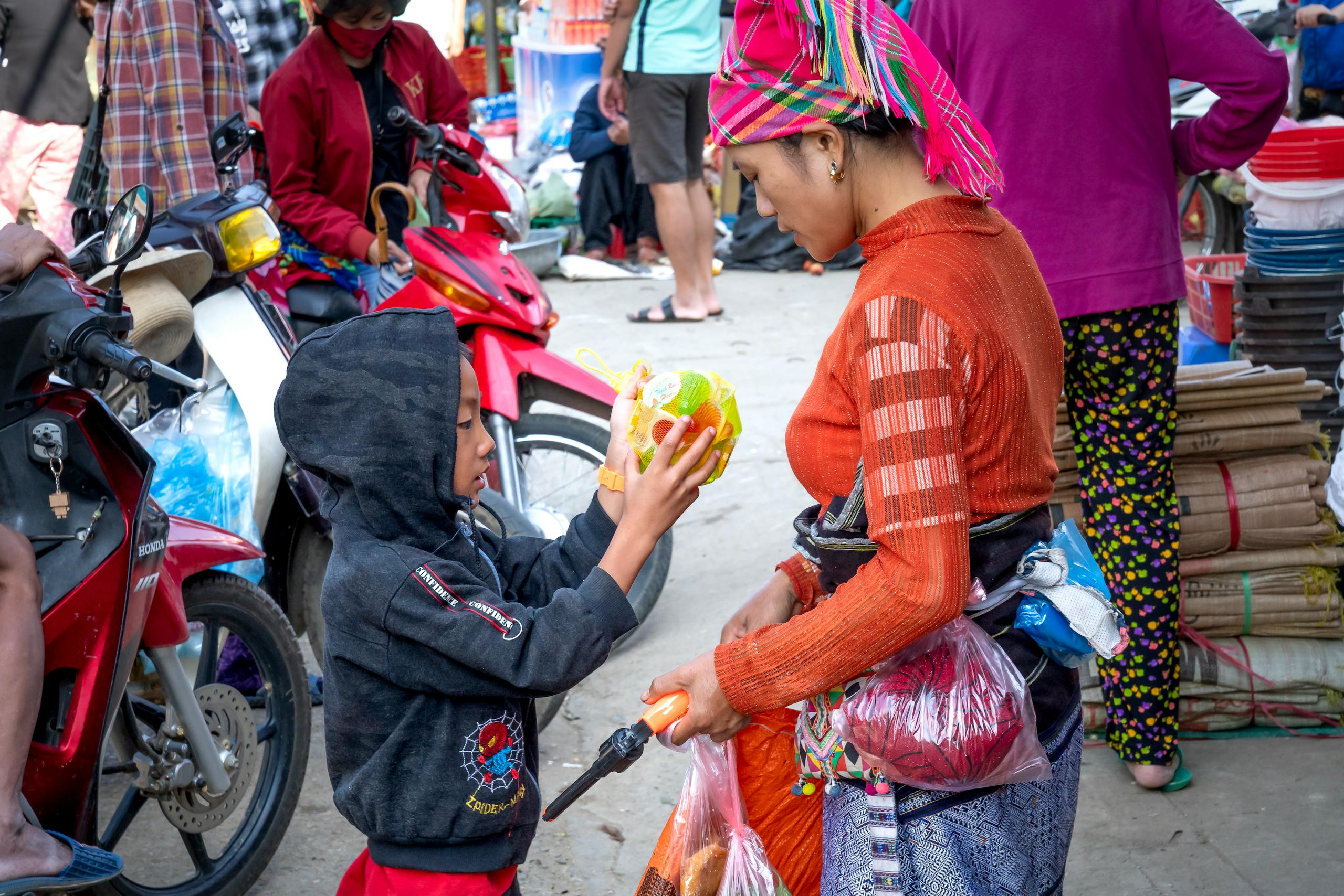 Asian mom in traditional wear against boy with decorative ball on pavement with unrecognizable people in bazaar