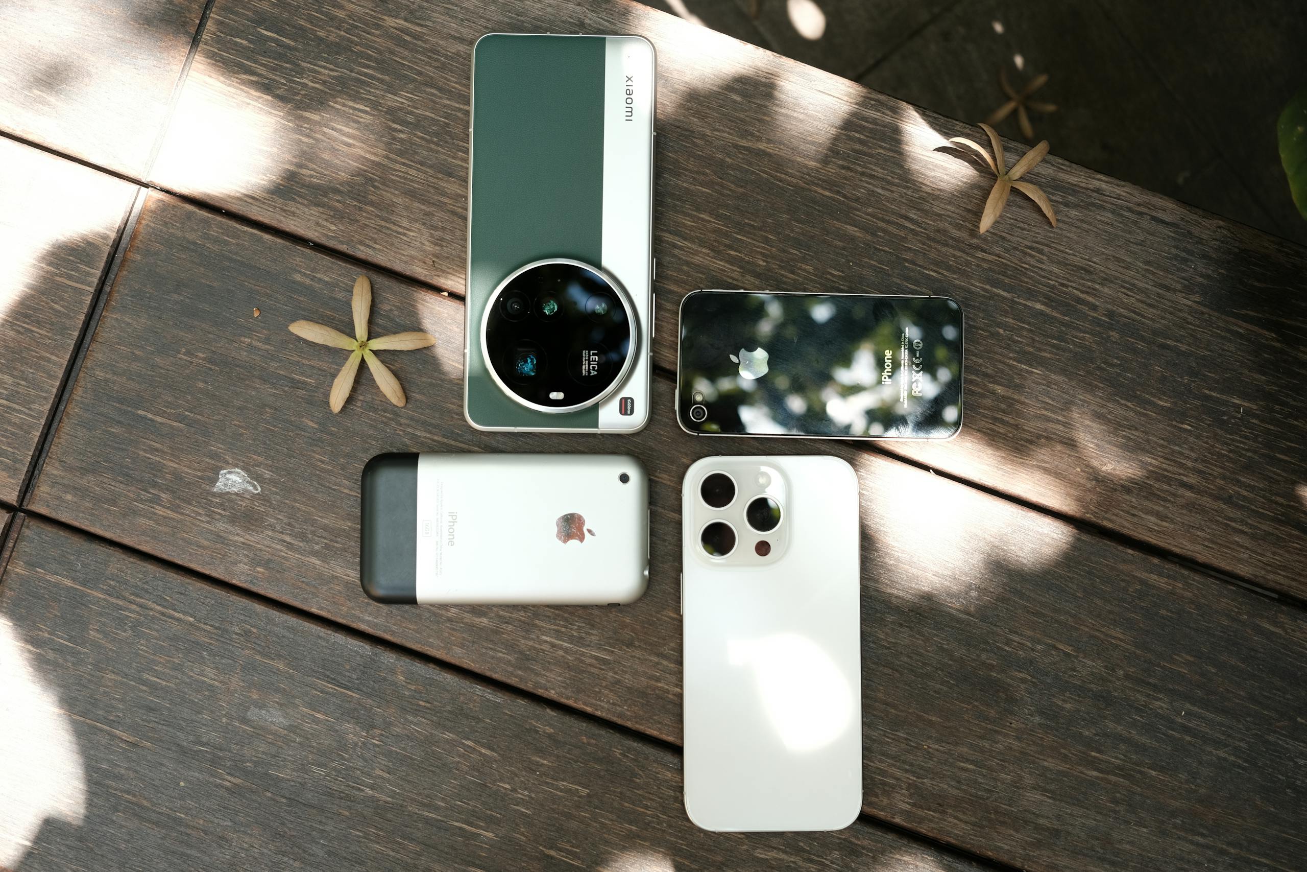 Assorted smartphones displayed outdoors on a wooden table under dappled sunlight.
