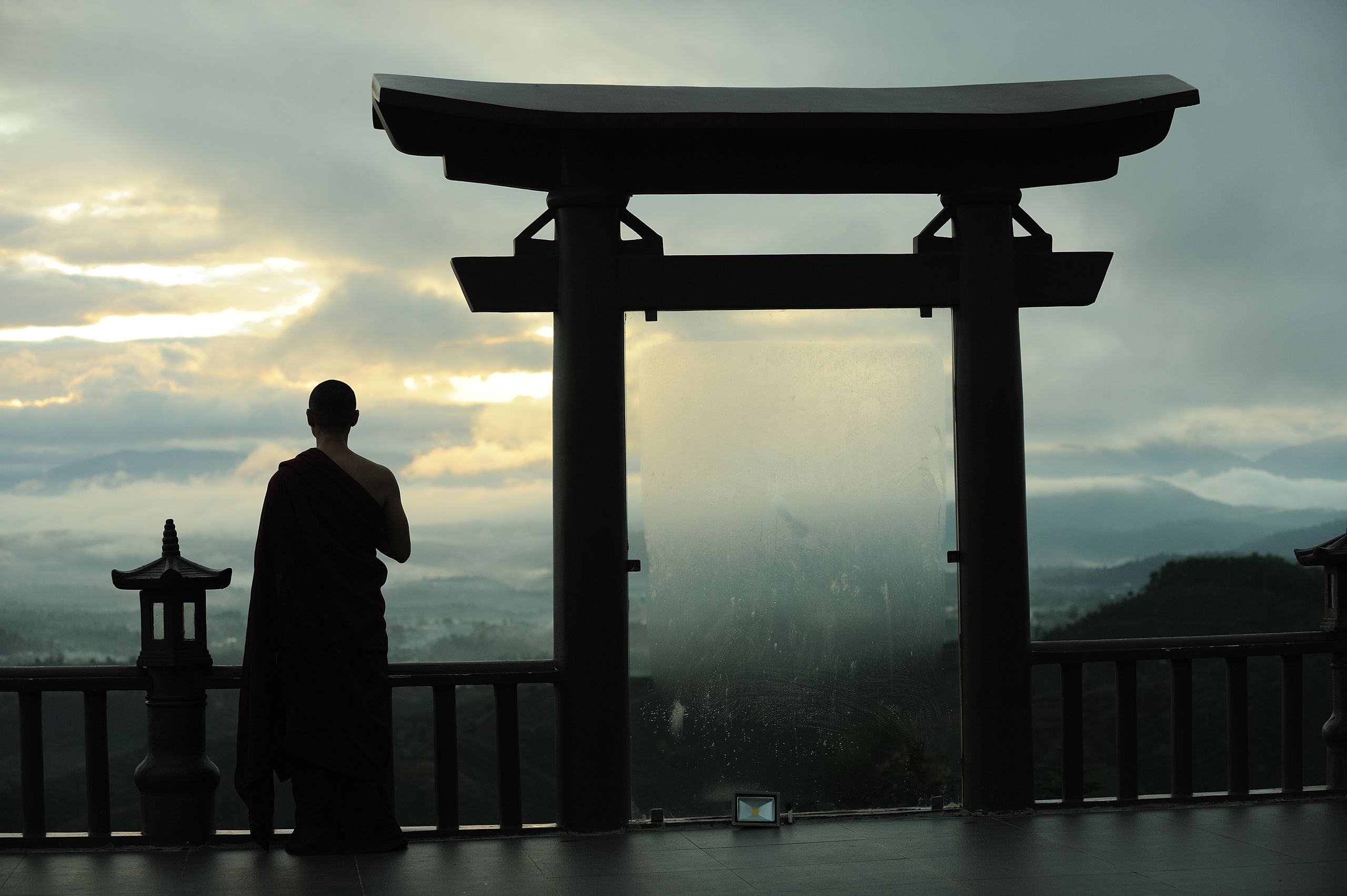 Back view full length anonymous monk in traditional robe standing near Torii Gate at entrance of Buddhist shrine and admiring tranquil scenery of evening exotic nature