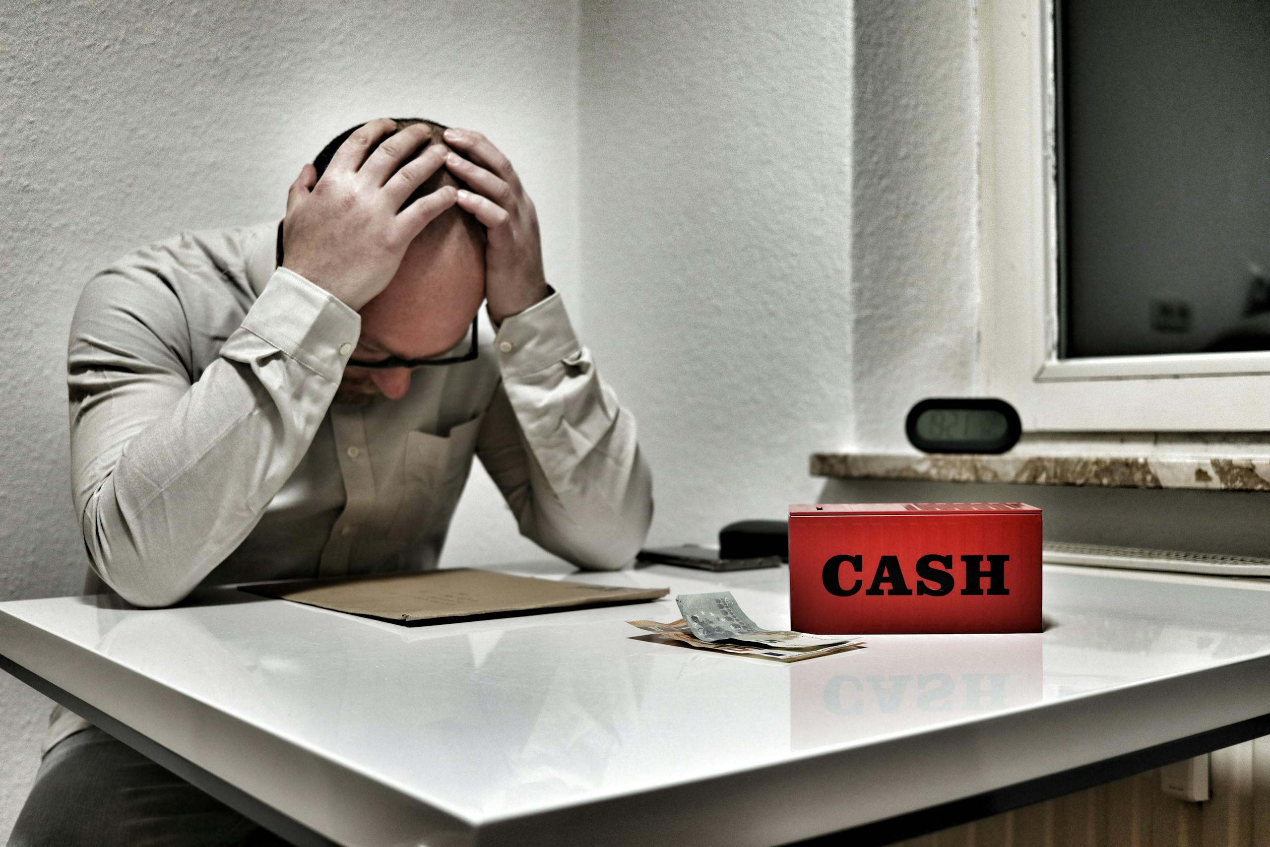 Bald man holding head in despair at desk with cashbox and money. Indoor office stress concept.
