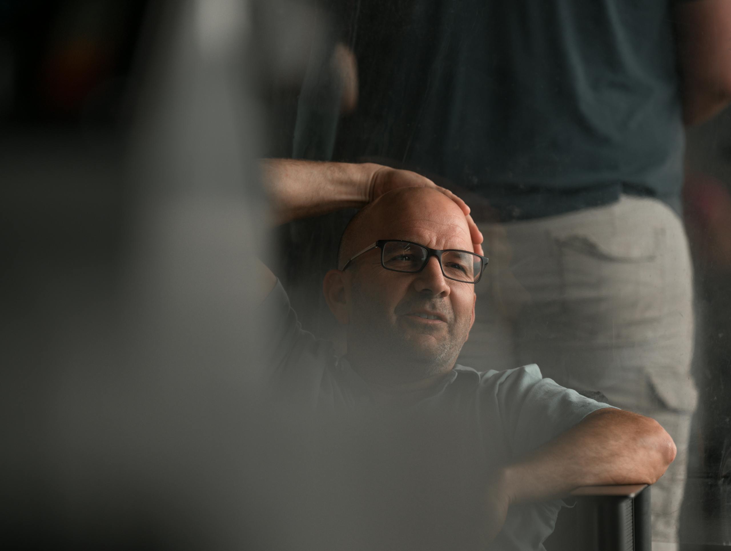 Bald man in glasses relaxing indoors with his hand on his head, thoughtful expression.