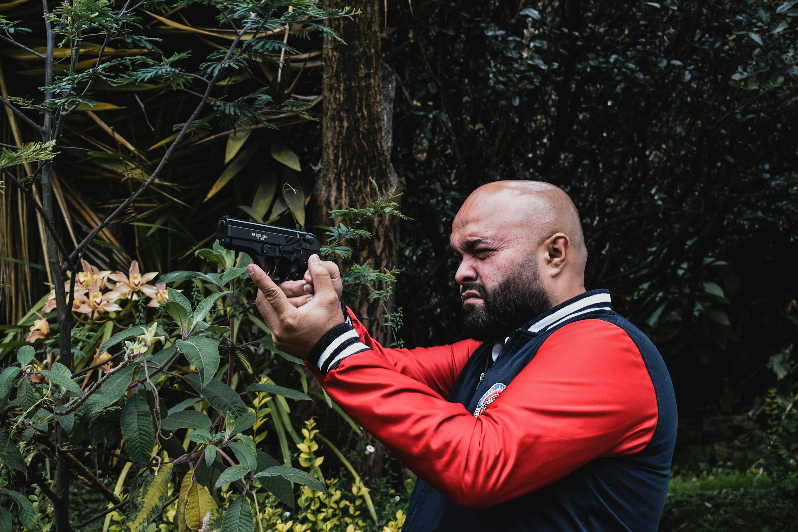 Bearded man in a red jacket aiming a black handgun amidst lush green foliage.
