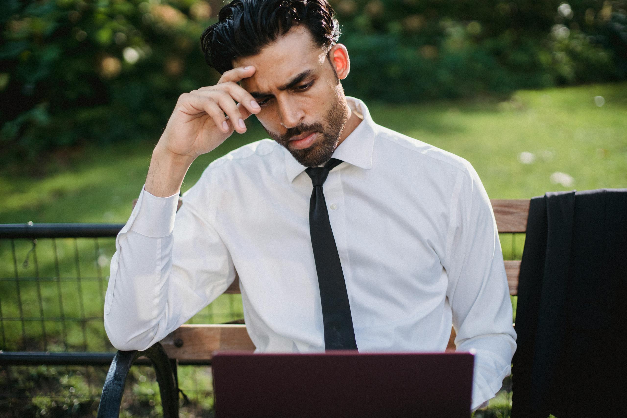 Bearded man in a white shirt and tie working on a laptop on a park bench.
