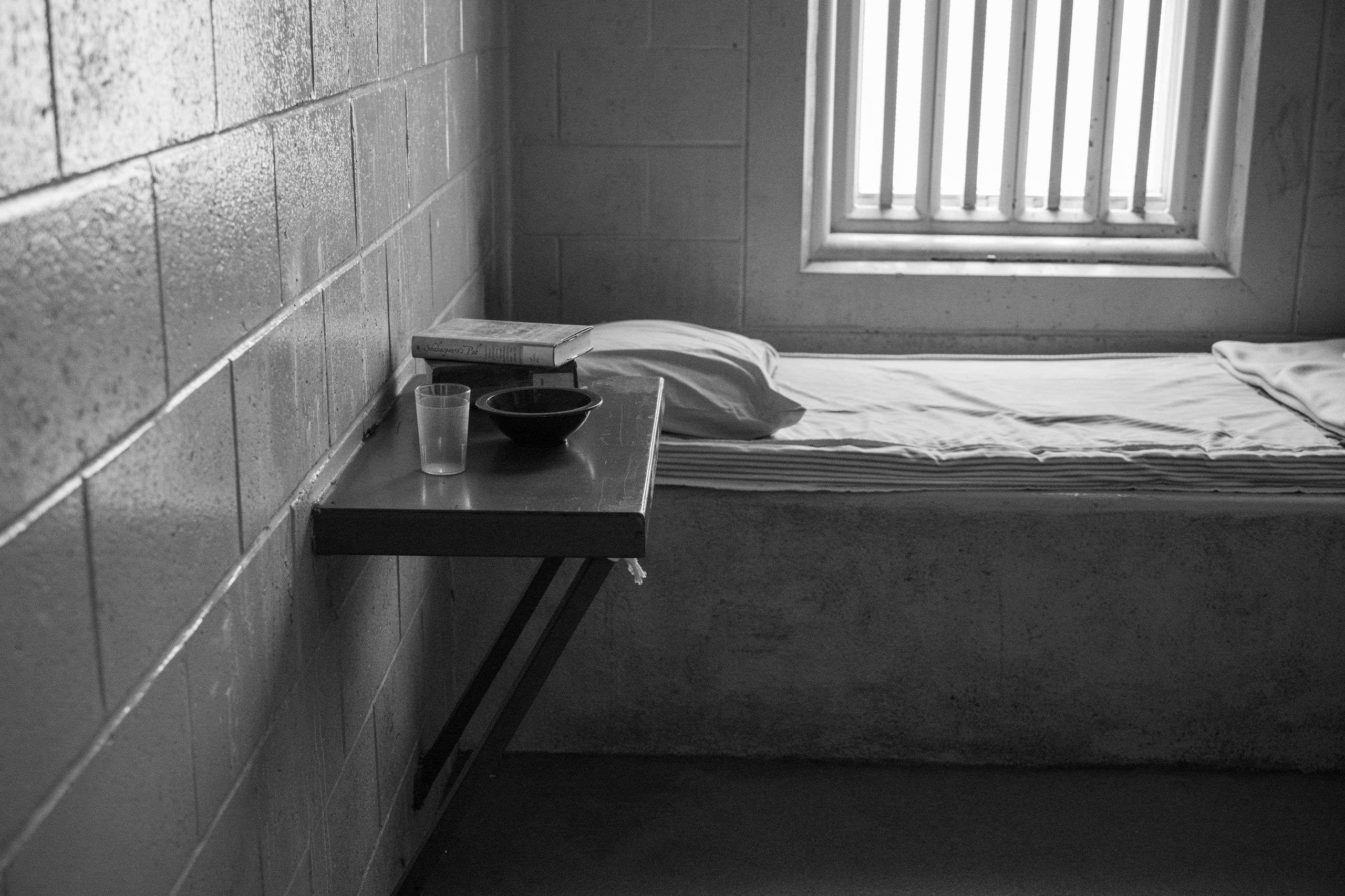 Black and white image of a minimalist prison cell with a bed, table, and window.